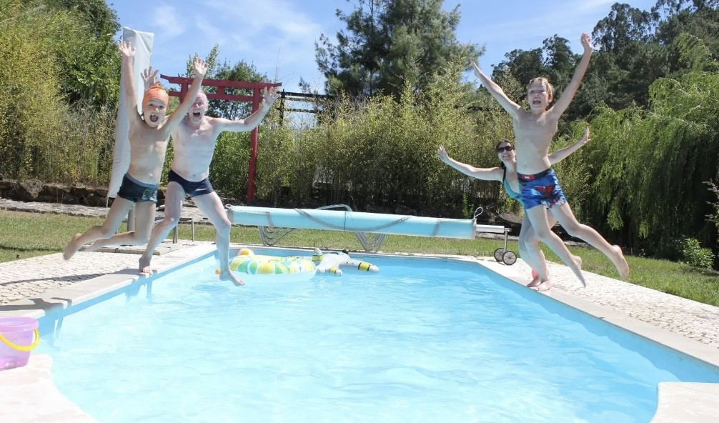 Four children jumping into a rectangular outdoor pool surrounded by grass and trees on a sunny day.