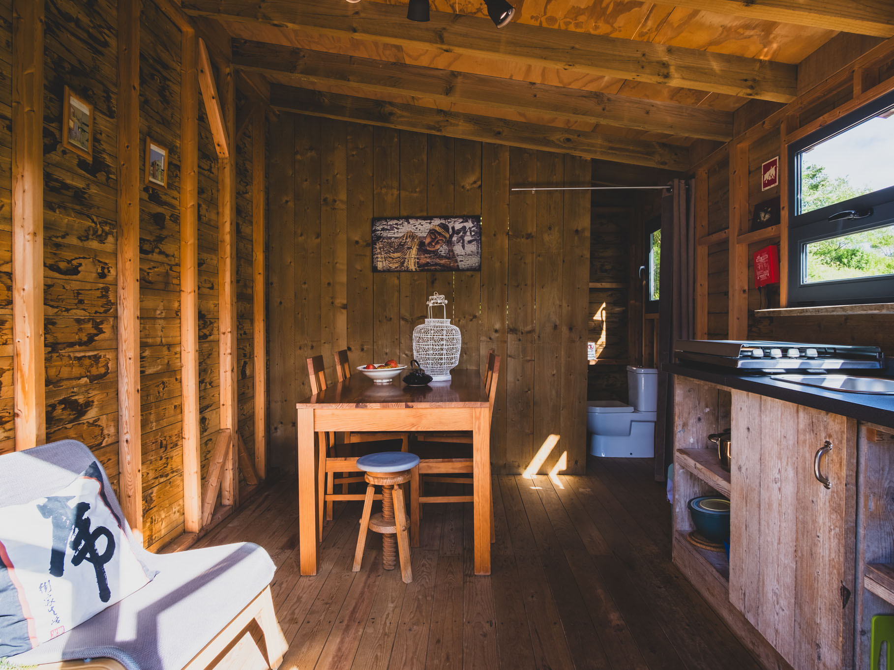 Compact wooden interior with dining table, chairs, kitchen counter, window, and visible bathroom in the background.