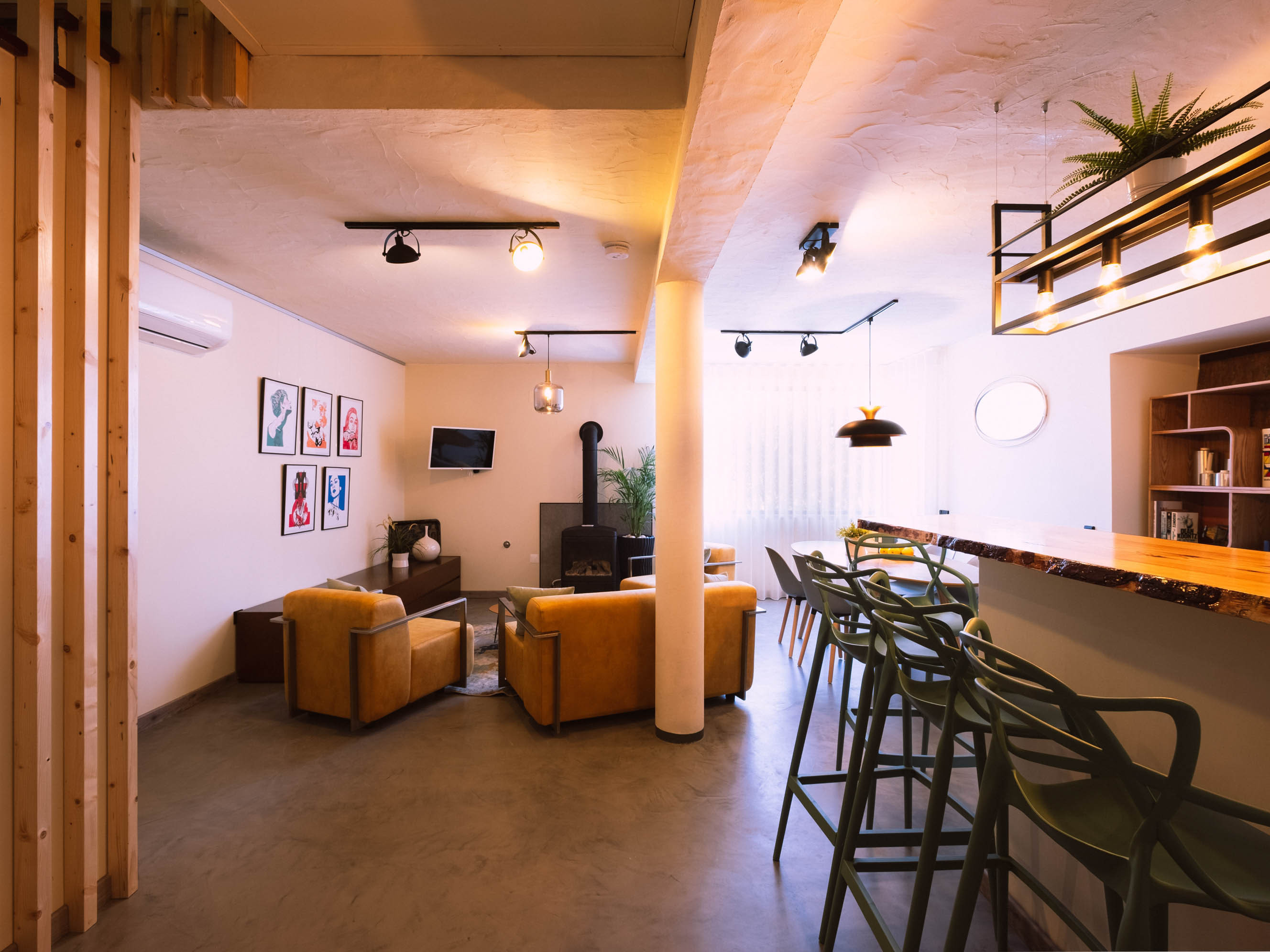 Living room with mustard armchairs, wood stove, wall TV, bar counter with stools, and dining table with chairs.