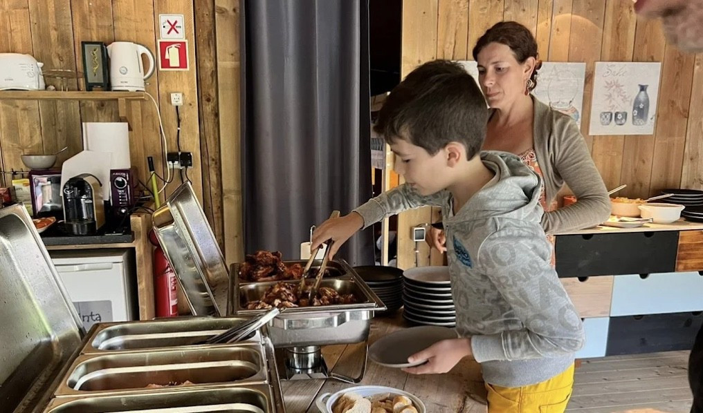 A boy serving food from a buffet table with a woman standing behind him in a wooden room.