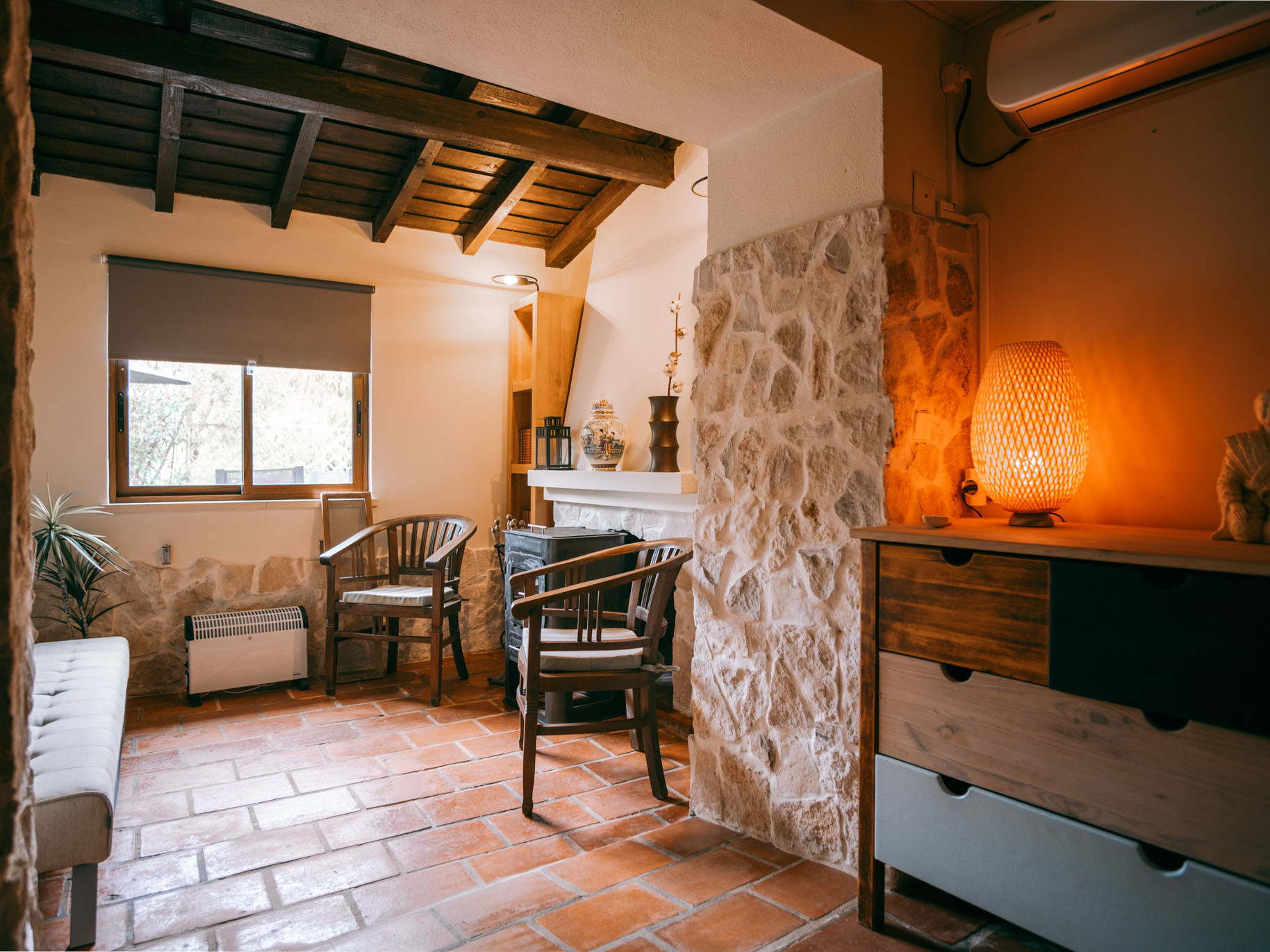 Cozy living room with wooden beams, terracotta floor, two wooden chairs, a window, and a glowing woven lamp.