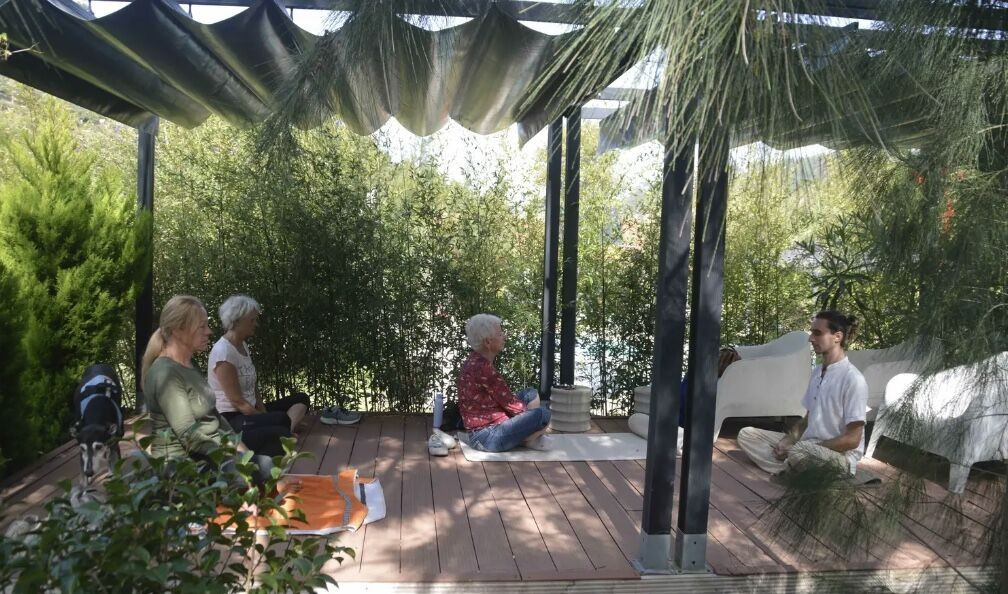 Four people sitting cross-legged on a wooden deck under a shaded pergola surrounded by greenery.
