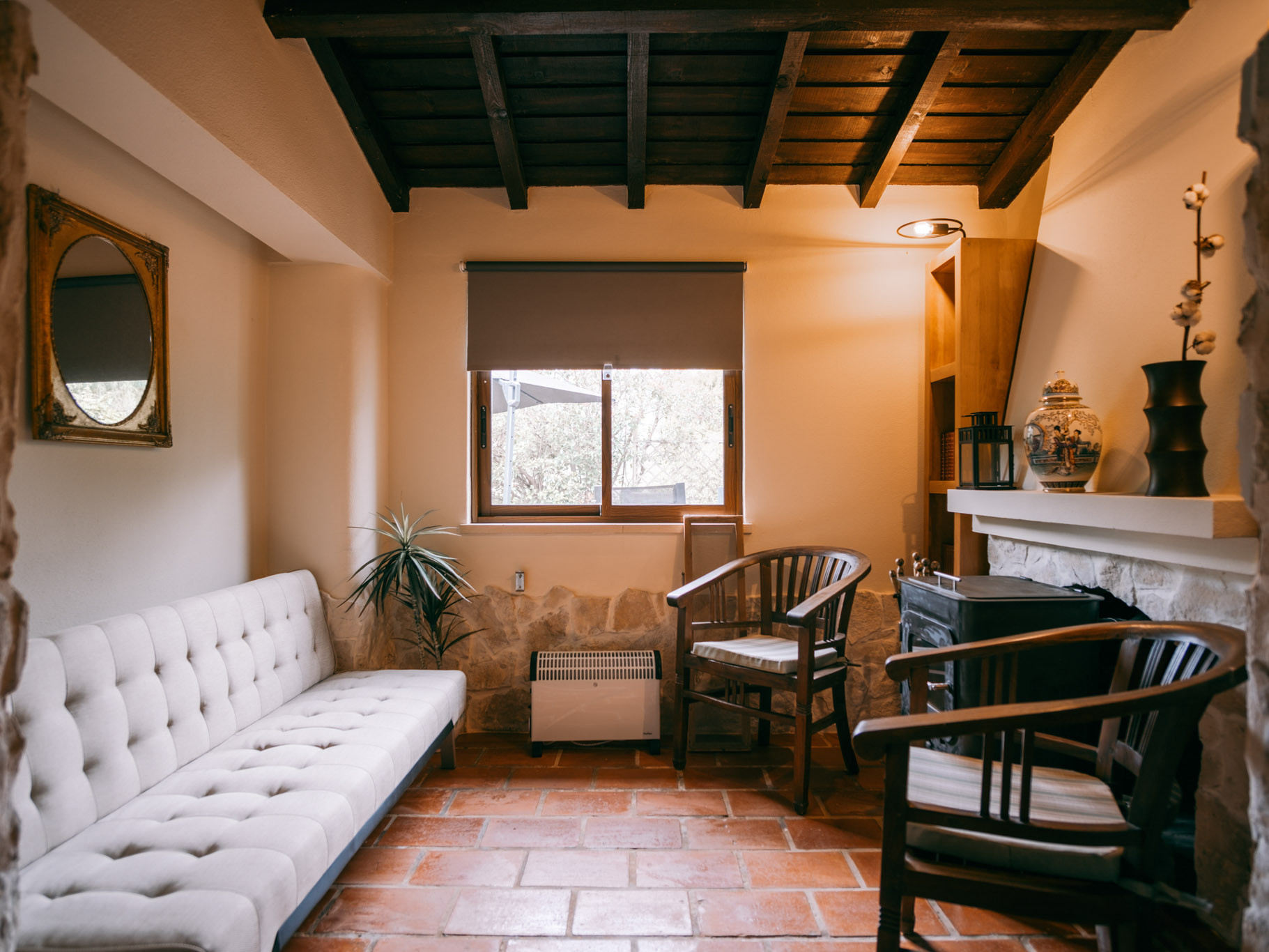 Cozy room with white tufted sofa, two wooden chairs, potted plant, window with roller blind, and tiled floor.