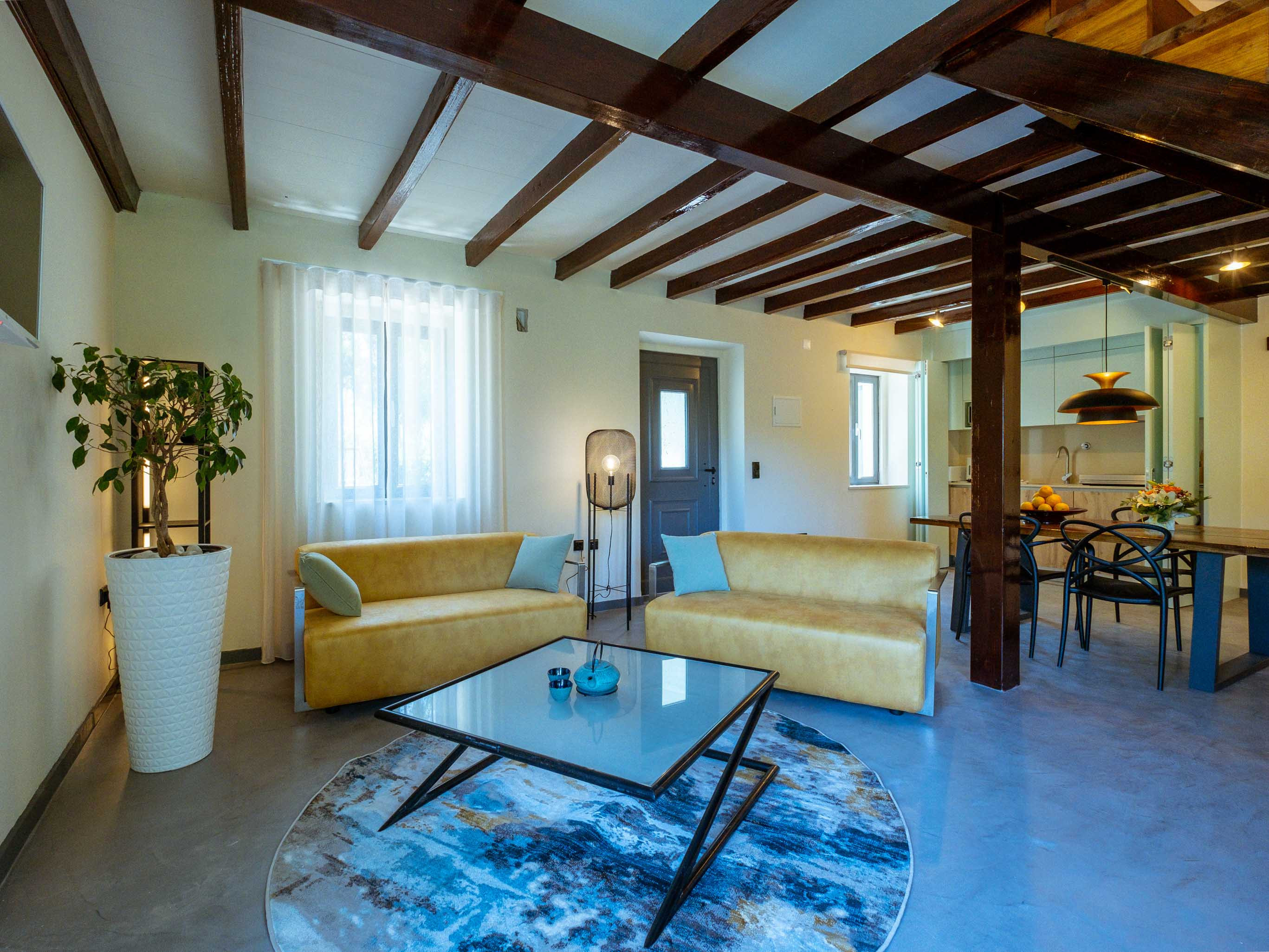 Living room with two beige sofas, glass coffee table on blue rug, wooden ceiling beams, and dining area.