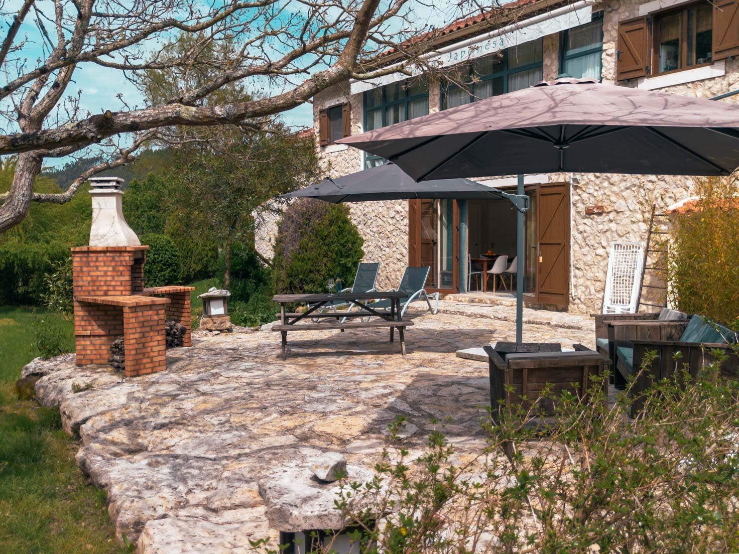 Stone patio with two large umbrellas, lounge chairs, wooden table, brick barbecue, and stone building facade.