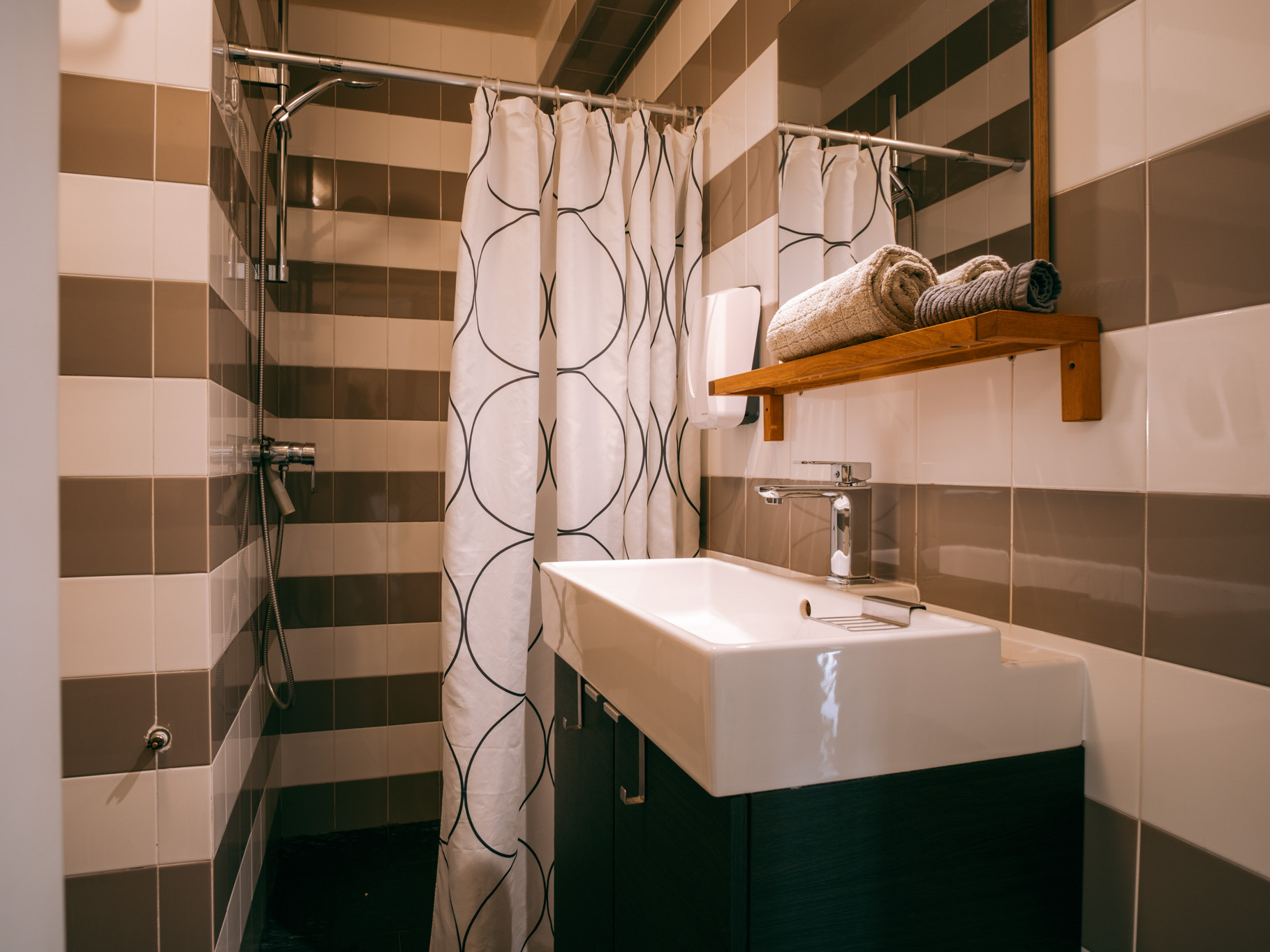 Bathroom sink and vanity beside a shower with patterned curtain, striped wall tiles, wooden shelf with towels.