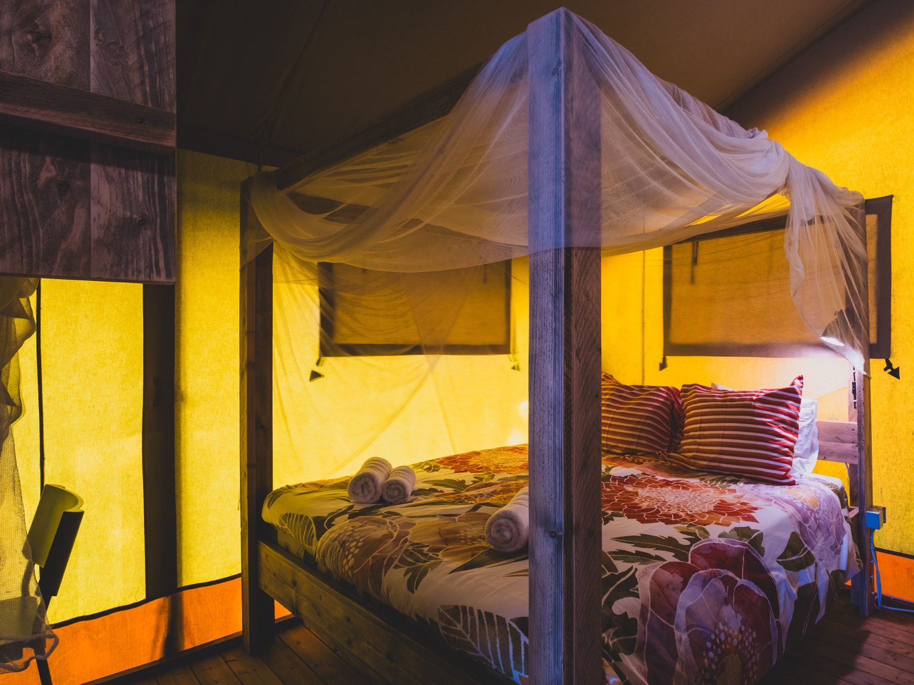 Double bed with floral bedding and striped pillows under light fabric canopy inside yellow tent room.