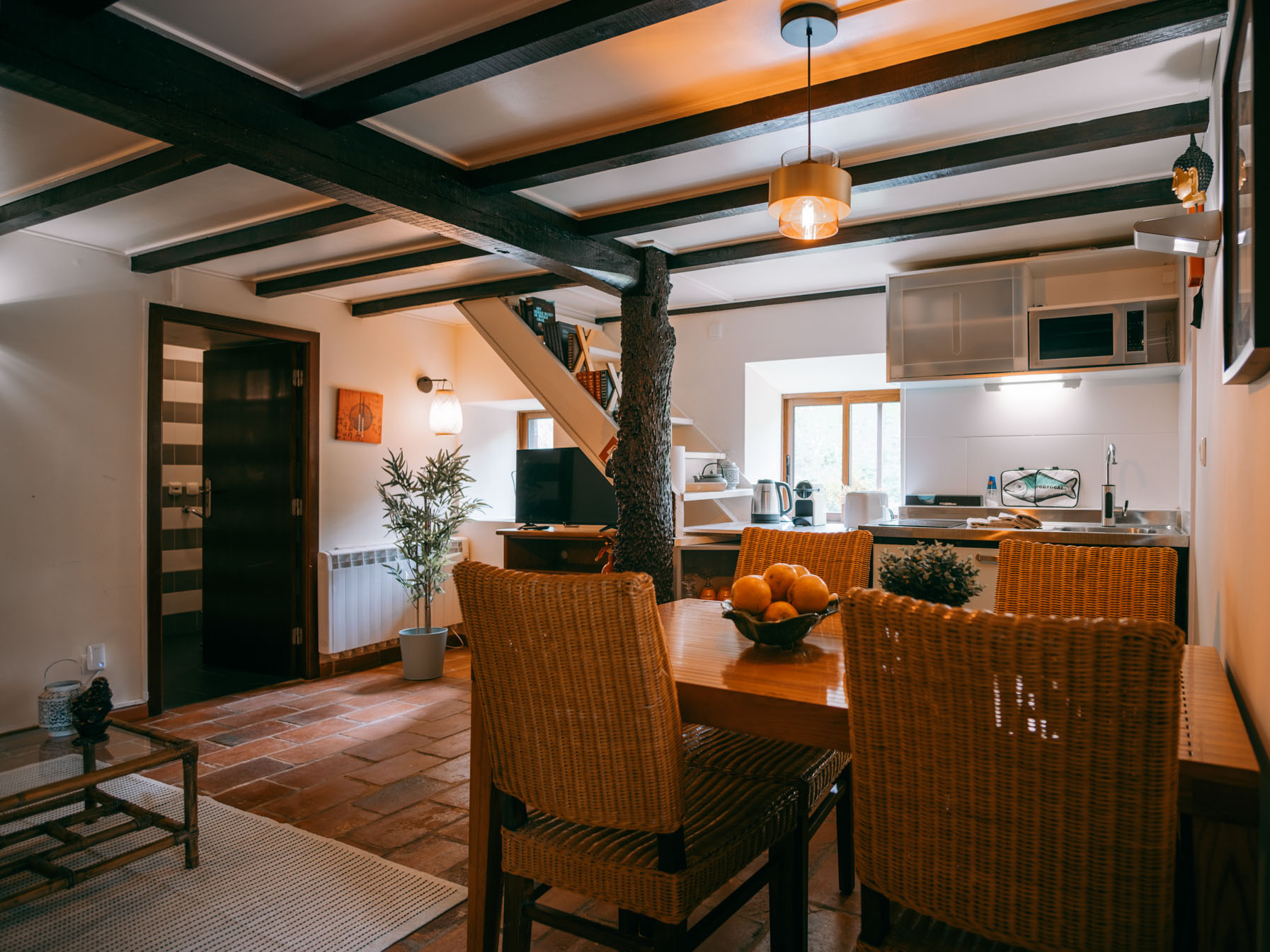 Dining table with woven chairs and fruit bowl in a cozy rustic kitchen-living room area.