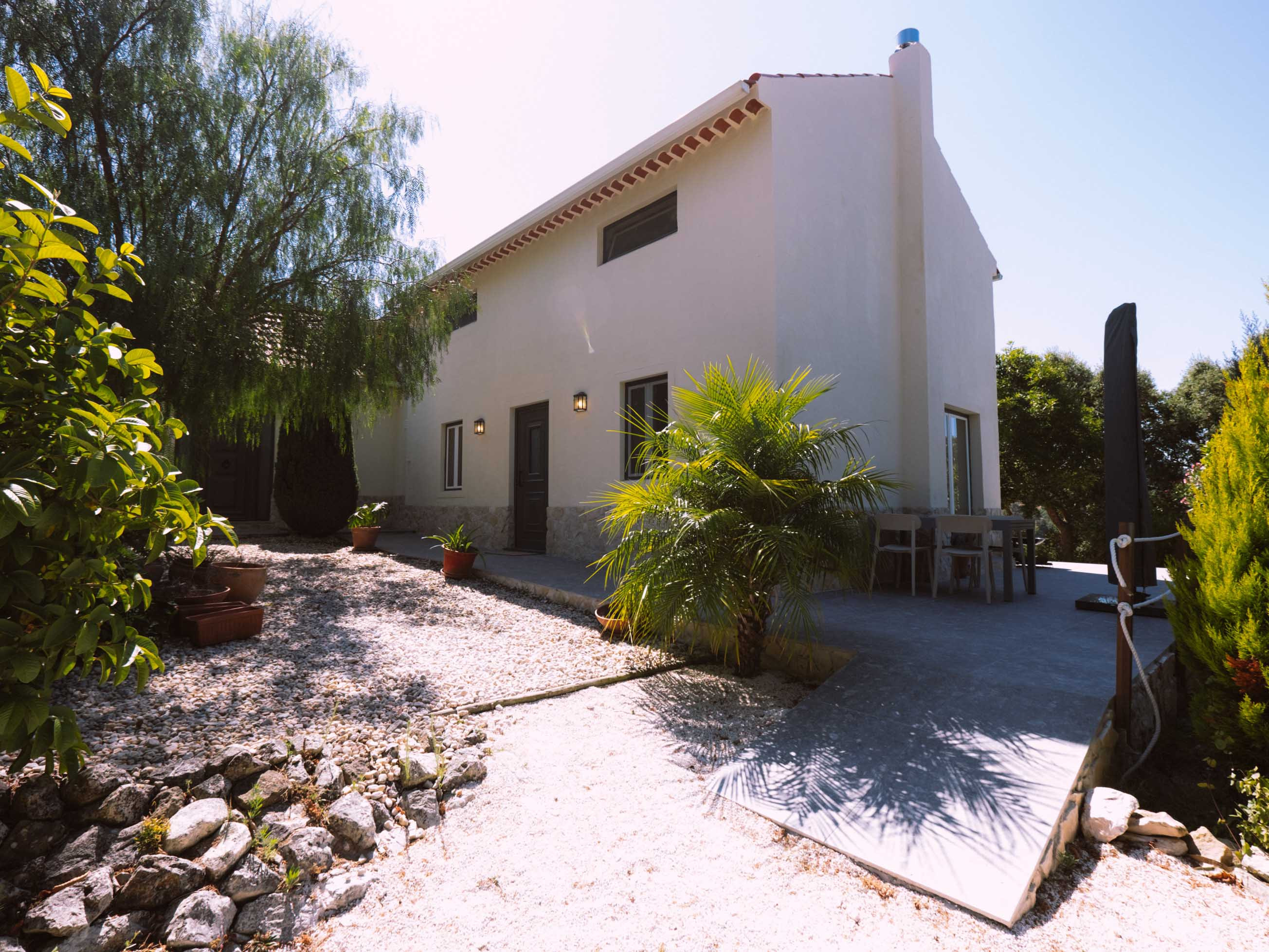 White two-story house with black door and windows, surrounded by gravel, plants, and shaded patio area.