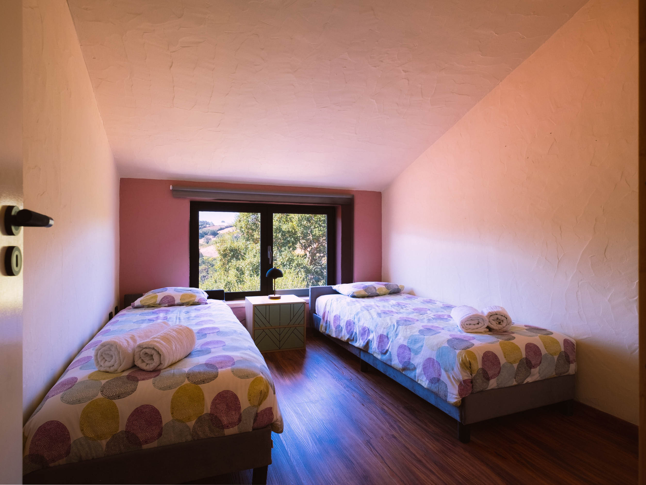 Bedroom with two single beds, colorful bedding, a nightstand with a lamp, and a window showing trees outside.
