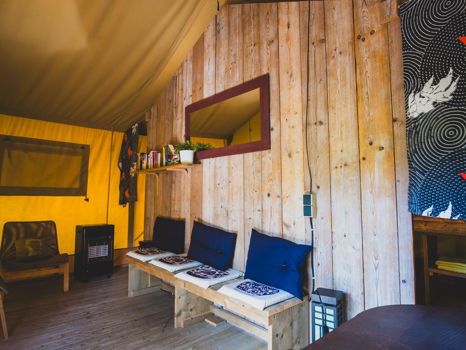 Indoor seating area with wooden bench, cushions, small shelf with plants and books, and wooden chair.
