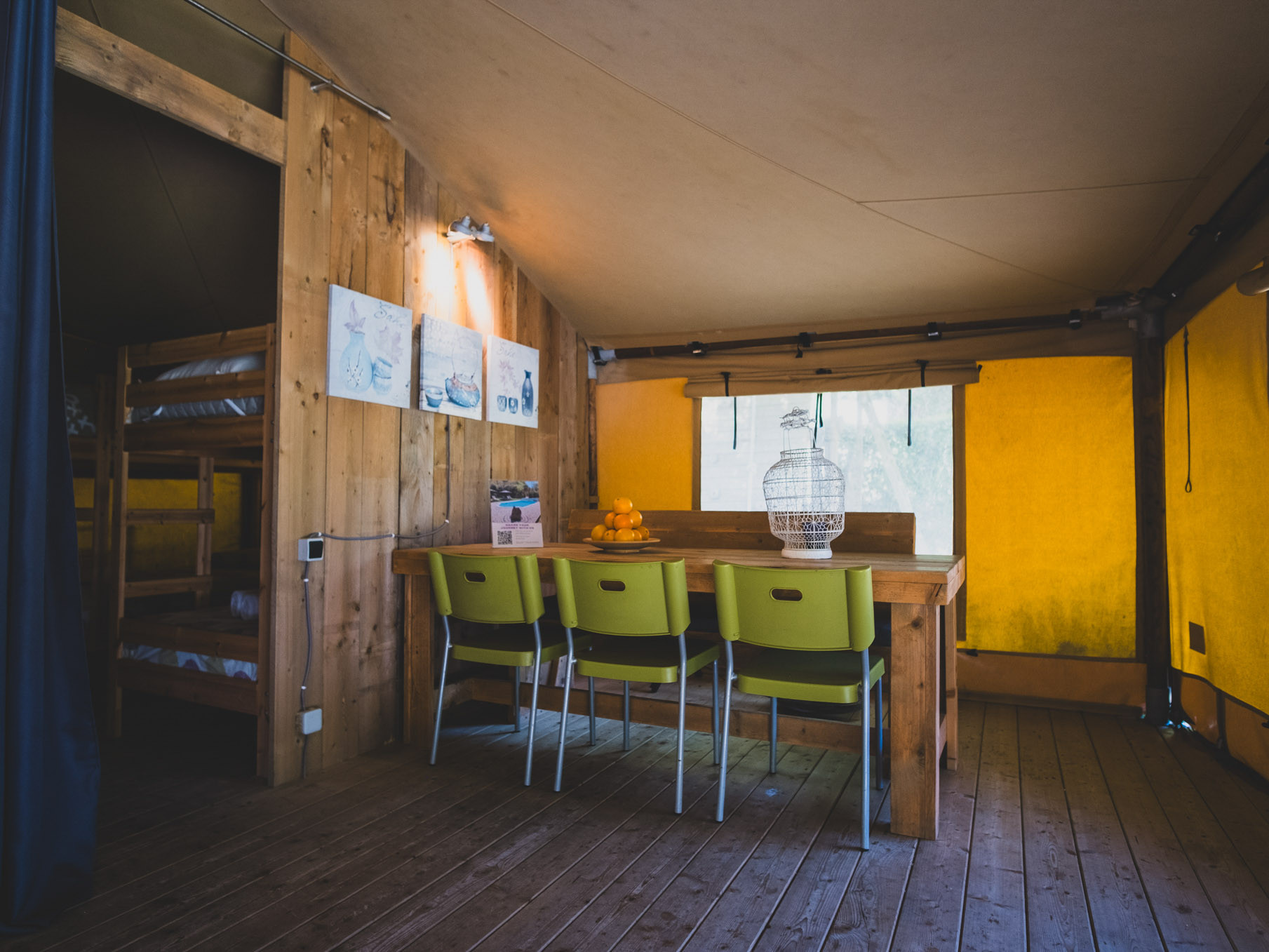 Wooden dining table with four green chairs inside a tent with bunk beds visible through doorway.