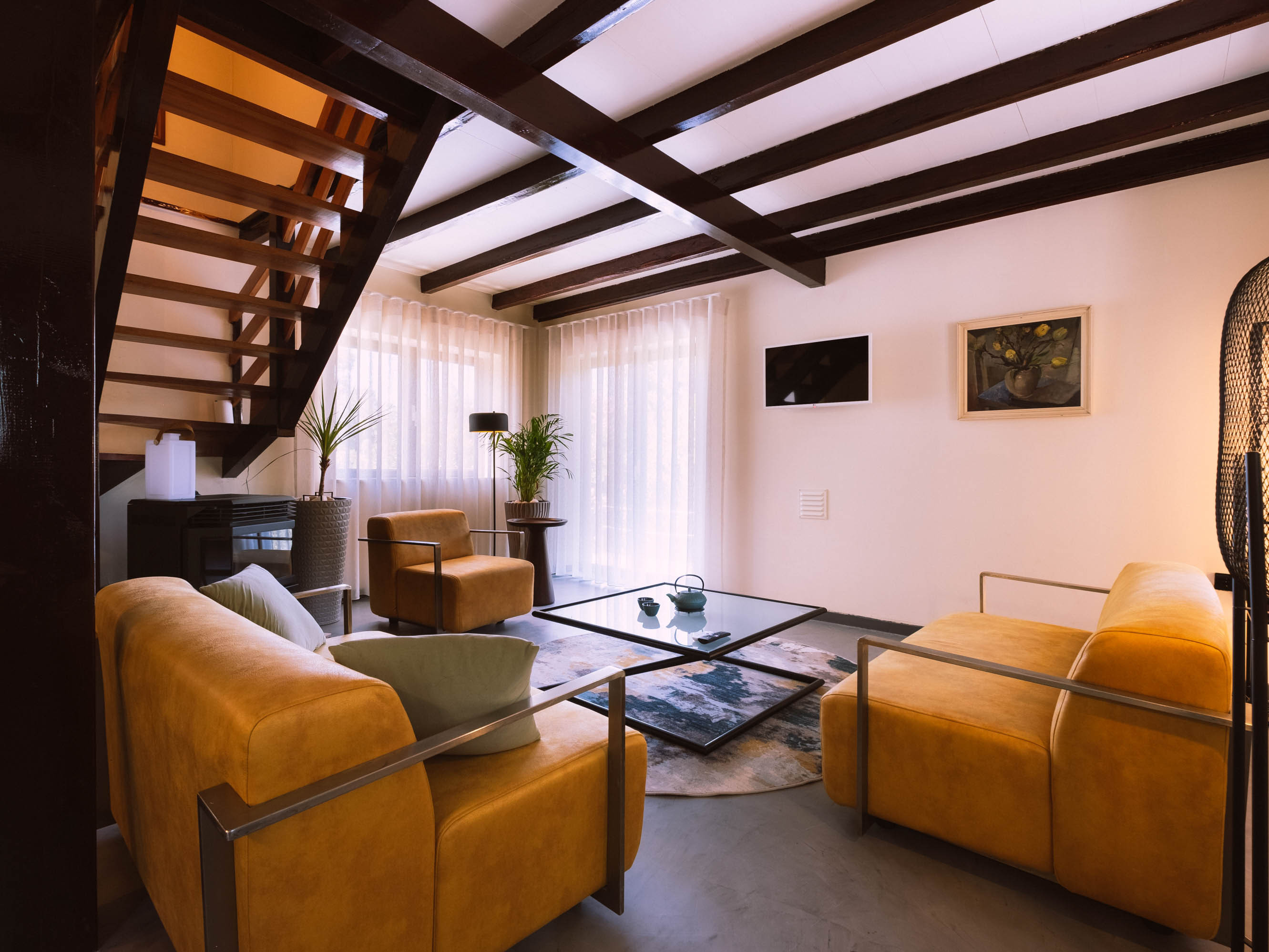 Living room with mustard yellow sofas, glass coffee table, plants, wooden staircase, and sheer curtains.