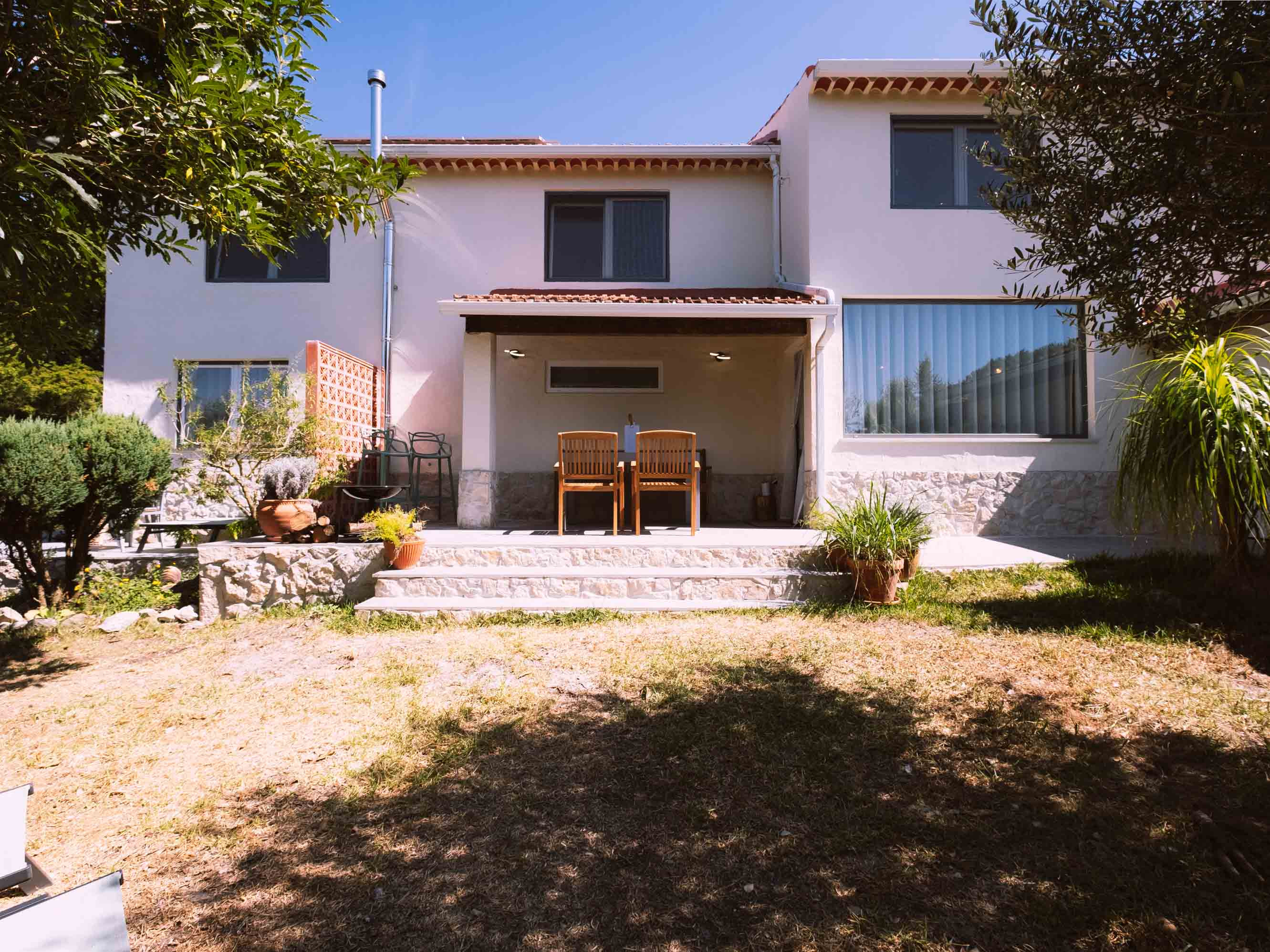 White two-story house with a small covered patio and two wooden chairs facing a garden.