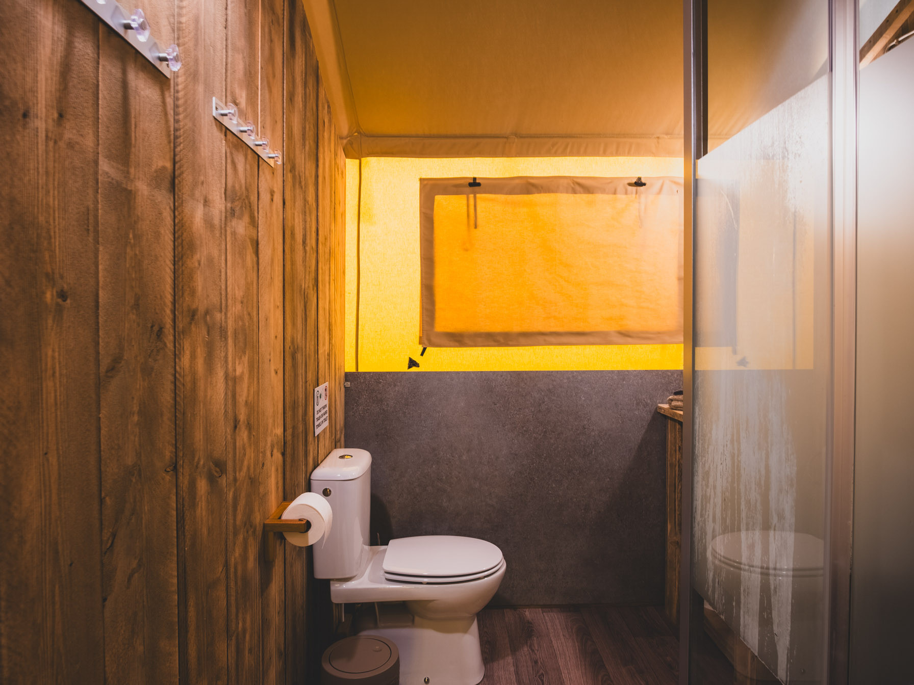 Bathroom with white toilet, wooden wall with hooks, frosted glass shower door, and yellow fabric window cover.