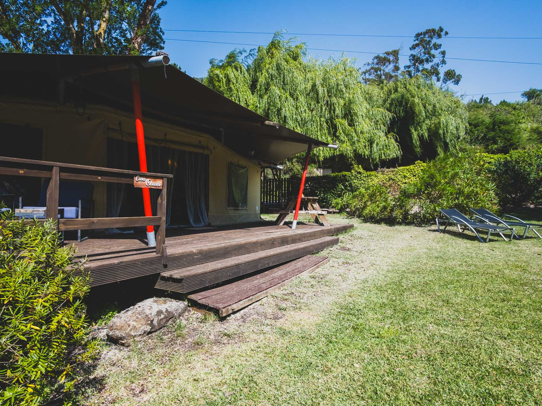 Wooden porch with red supports next to green lawn and lounge chairs under clear blue sky.