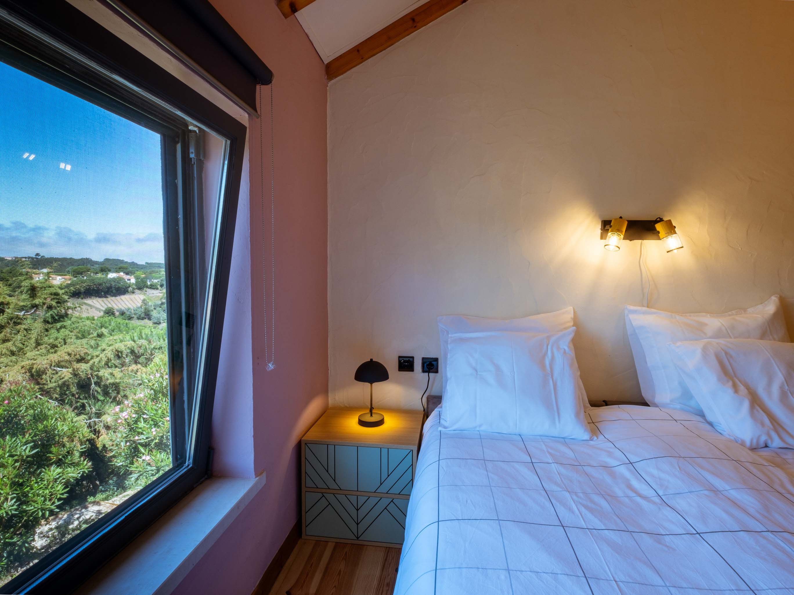 Bed with white linens next to a bedside table and window overlooking green landscape.