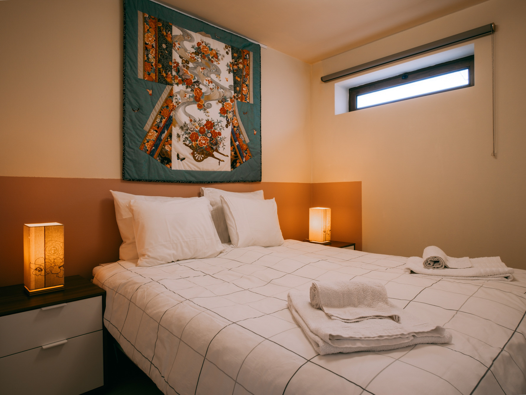 Double bed with white linens, folded towels, bedside lamps, decorative textile hanging and small high window.