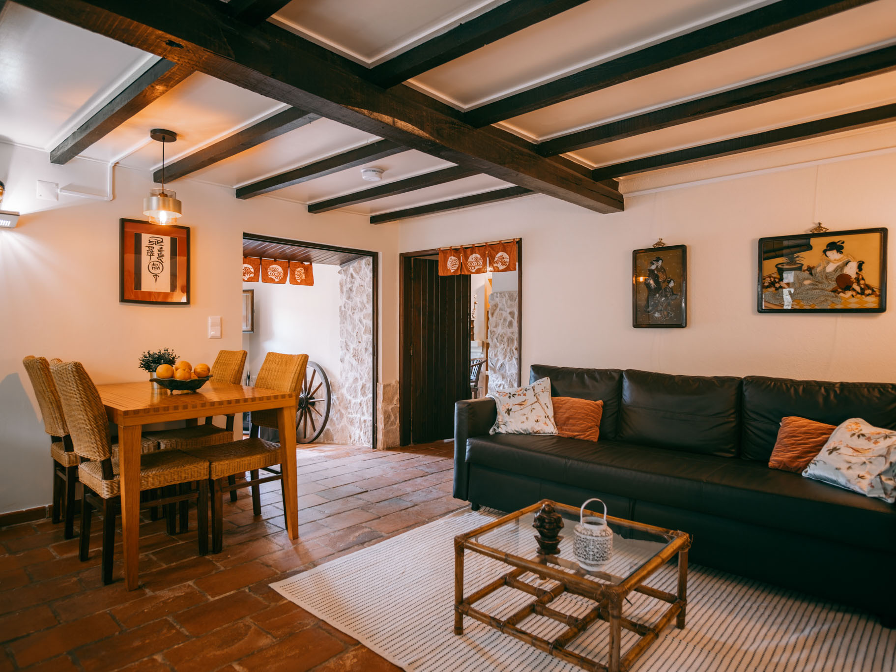 Cozy living and dining area with wicker chairs, wooden table, black sofa, framed art, and exposed beams.