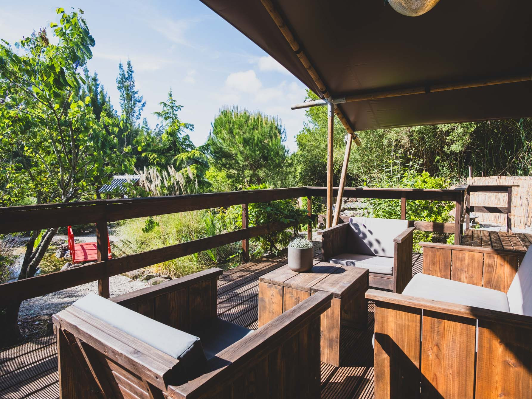 Wooden outdoor seating area with cushioned chairs and small table on a deck surrounded by greenery.