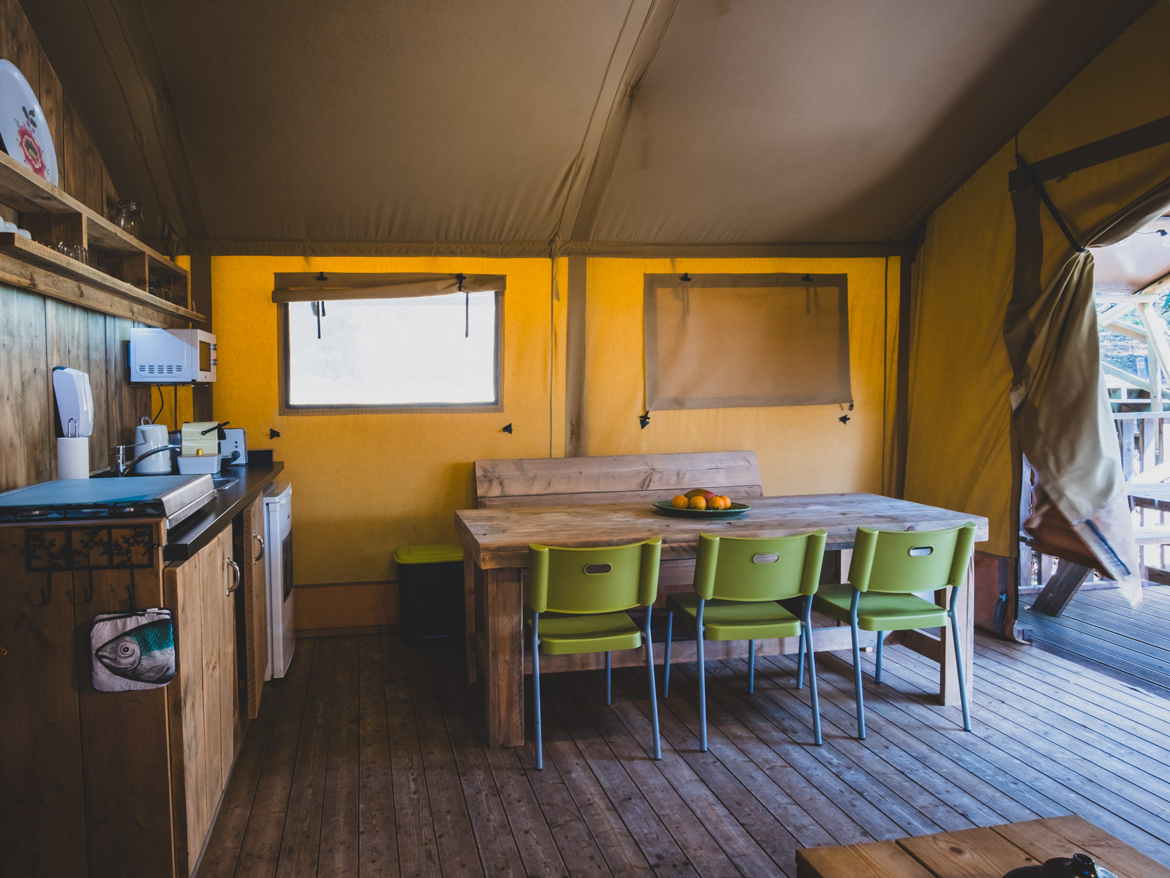 Interior of a tent with a wooden dining table, four green chairs, yellow walls, and a small kitchenette.