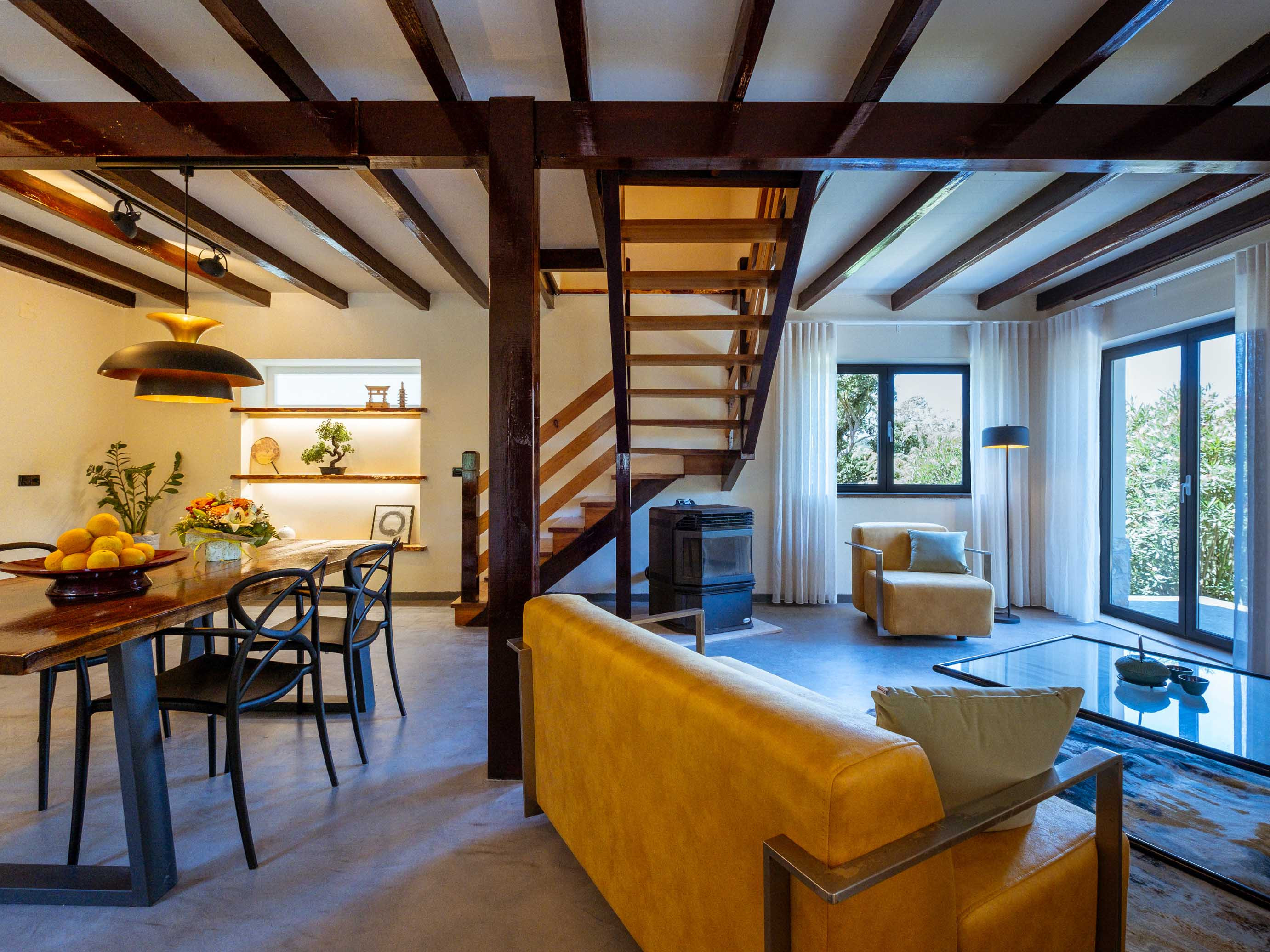 Living and dining area with yellow sofa, wooden dining table, chairs, and staircase under wooden ceiling beams.