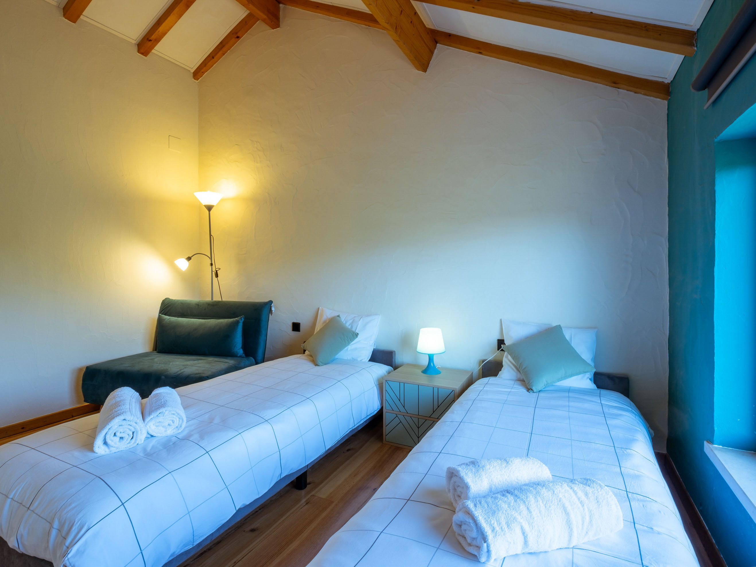 Bedroom with two single beds, folded towels, a nightstand with lamp, and a chair near a lamp-lit corner.