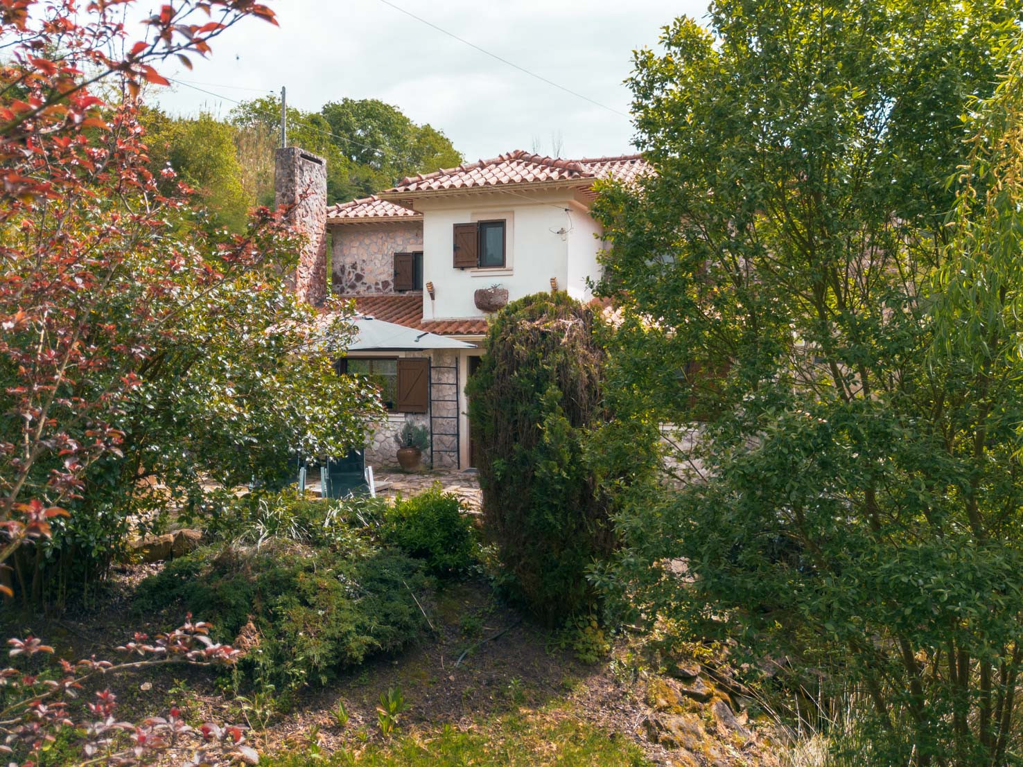 A two-story rustic house partially obscured by dense green and reddish foliage under a cloudy sky.
