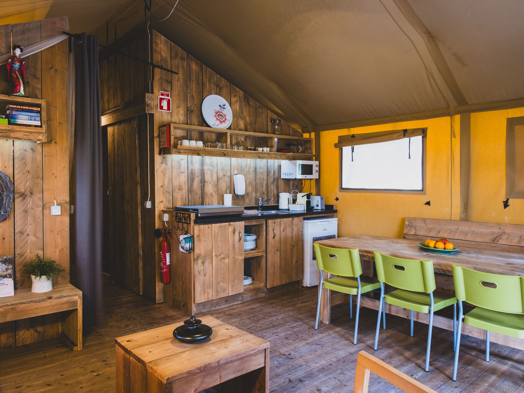 Interior of a wooden kitchen and dining area with green chairs around a wooden table and yellow walls.