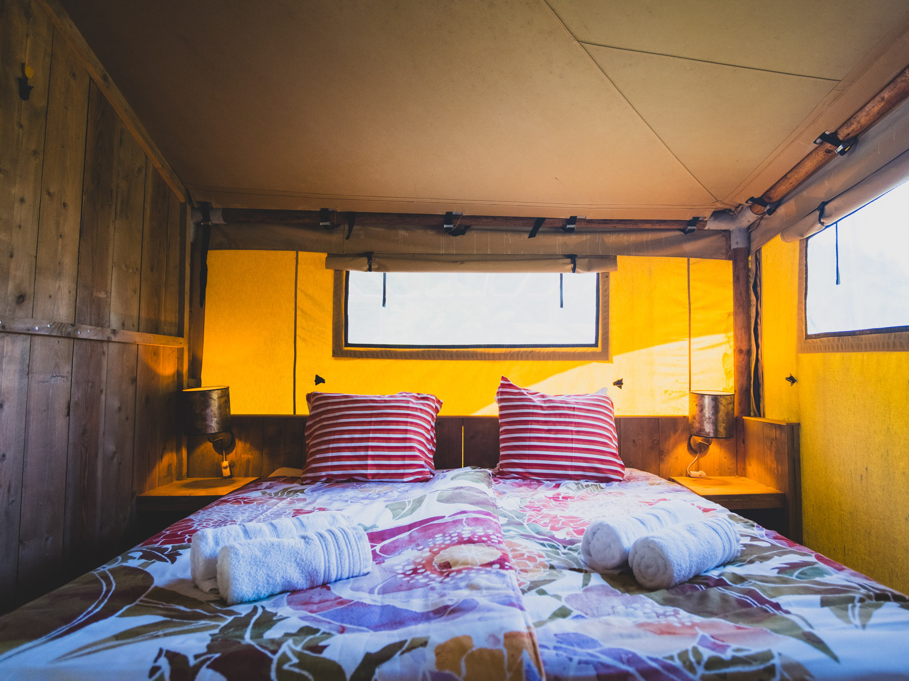 Bed with patterned blanket, two red-striped pillows, rolled towels, wooden side tables, and yellow canvas walls.