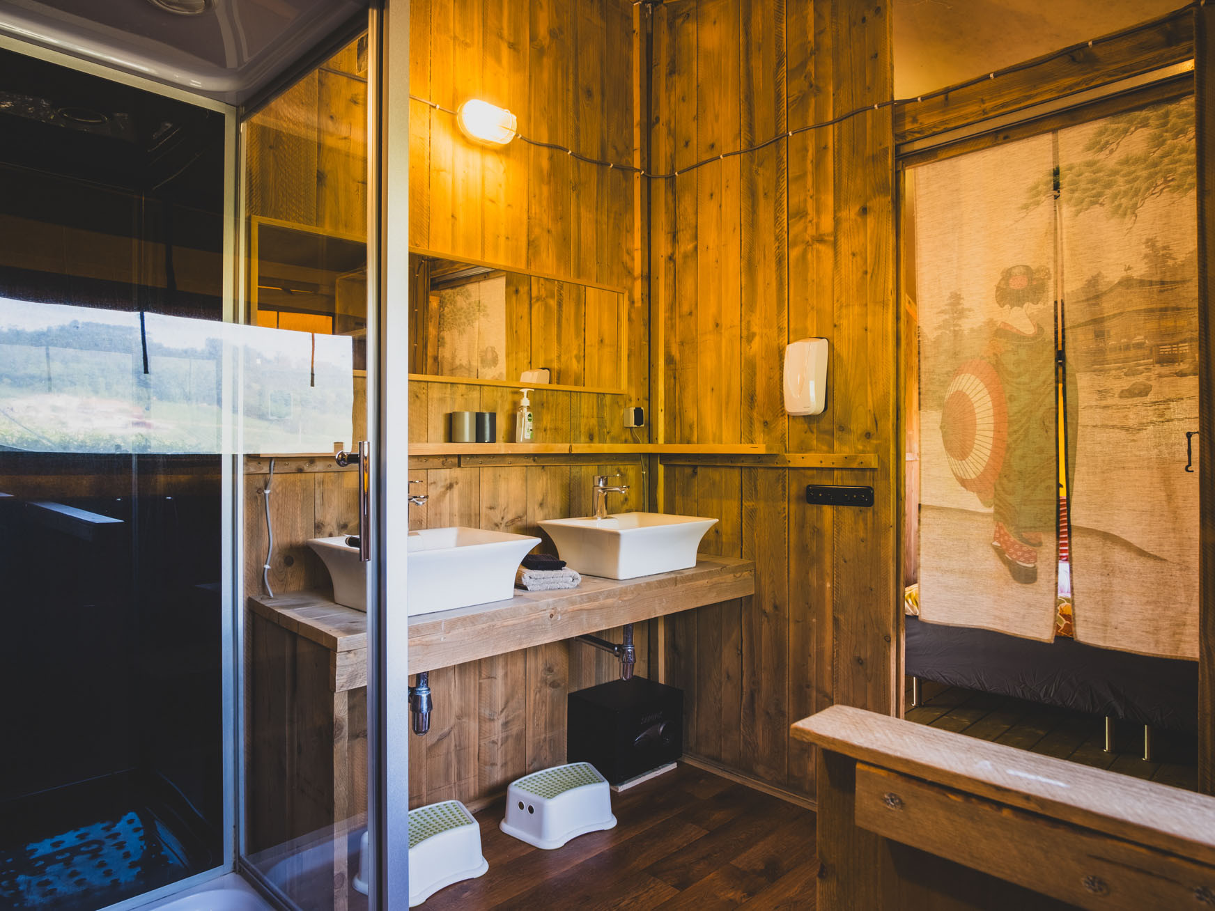 Wood-paneled bathroom with two vessel sinks, a mirror, small stools, and a decorative curtain featuring a figure.
