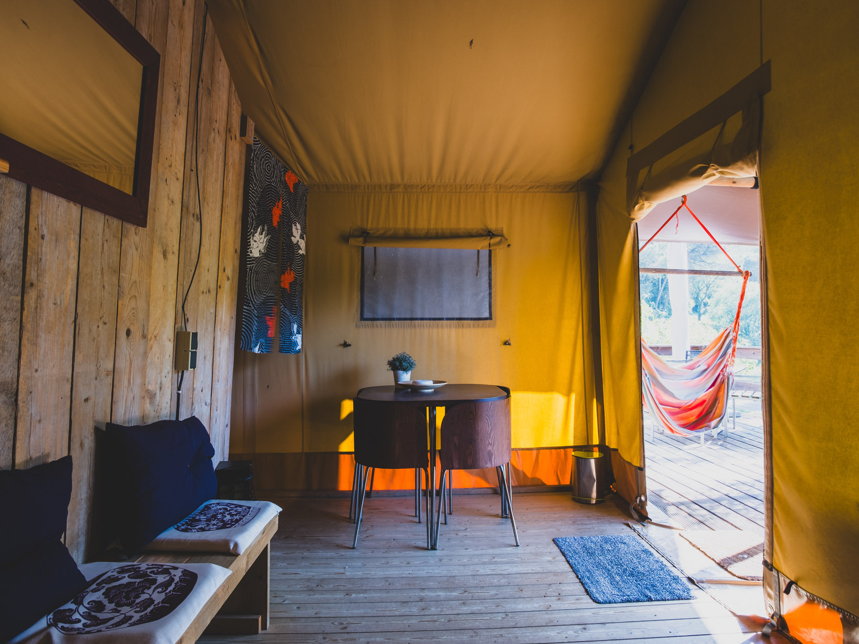 Interior of a wooden and fabric tent with seating bench, table with two chairs, and orange hammock outside.