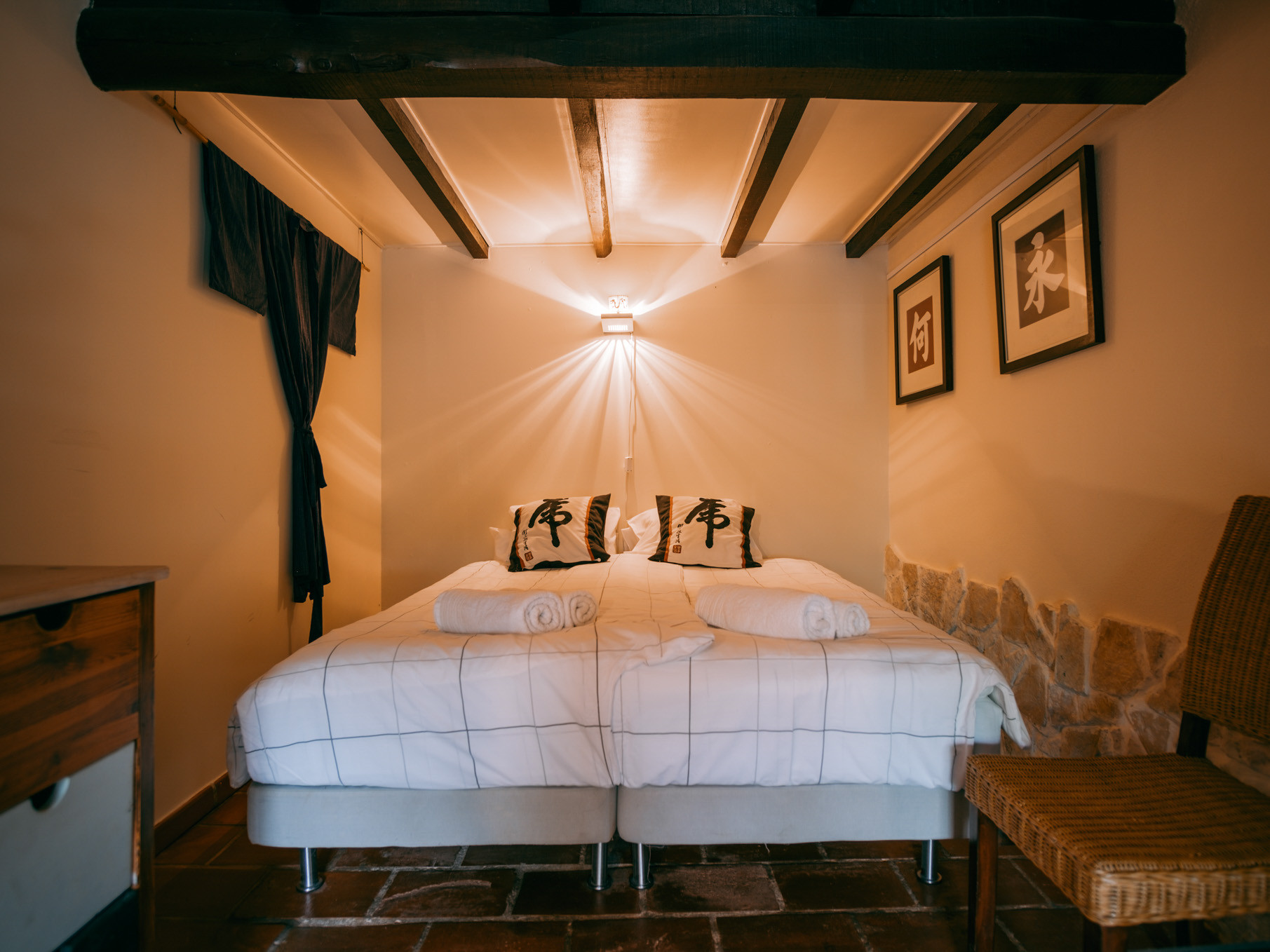 Cozy bedroom with two single beds, Japanese-themed pillows, wall art, warm lighting, and a wicker chair.