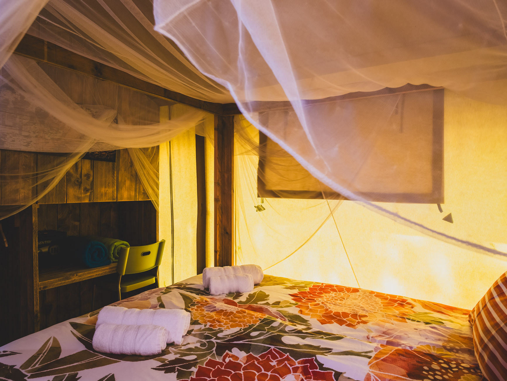 Bed with floral bedspread, rolled towels, and mosquito netting in warmly lit room with wooden furniture.