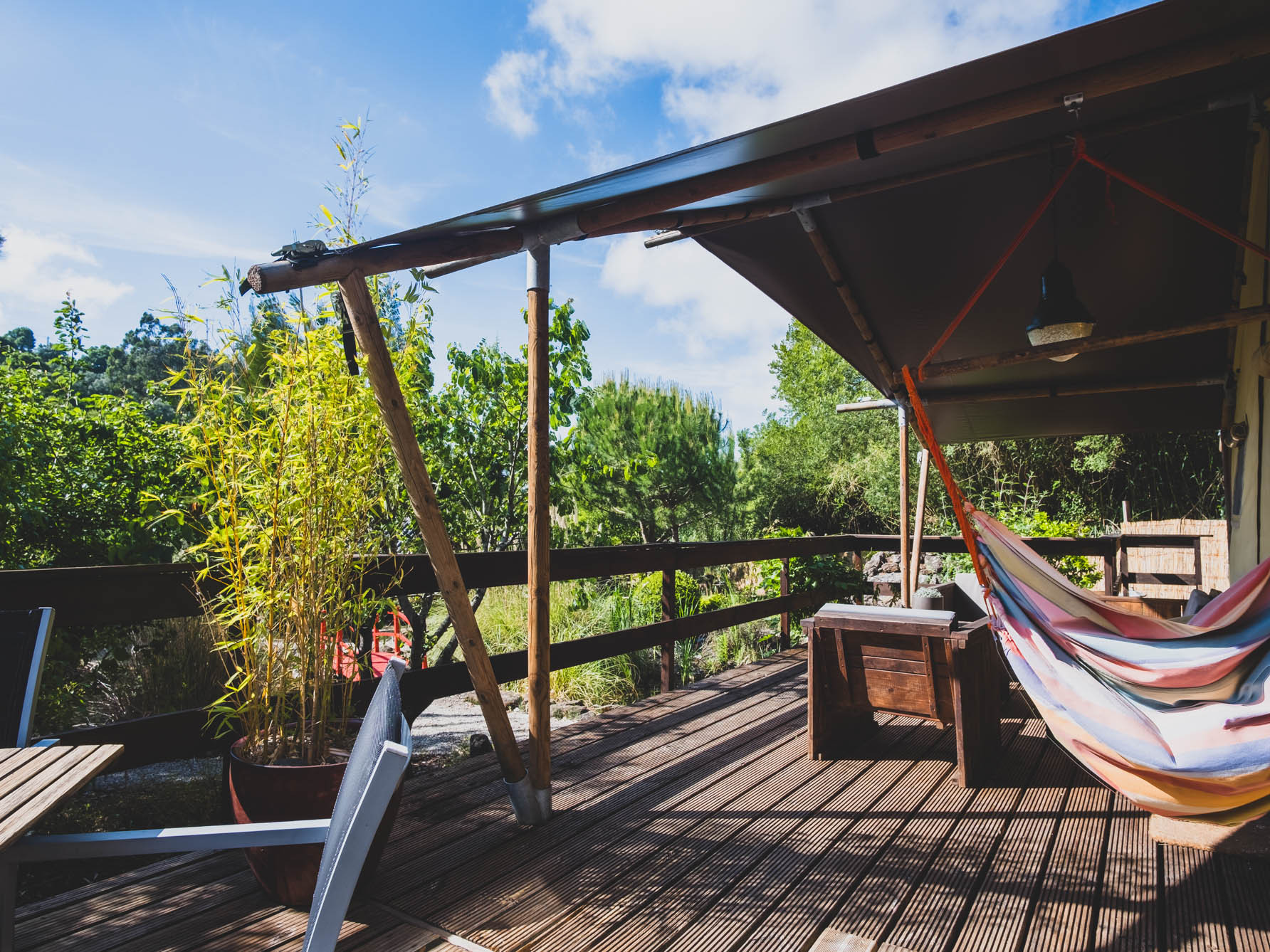 Wooden deck with chairs, table, hammock under shaded roof surrounded by green plants and trees.