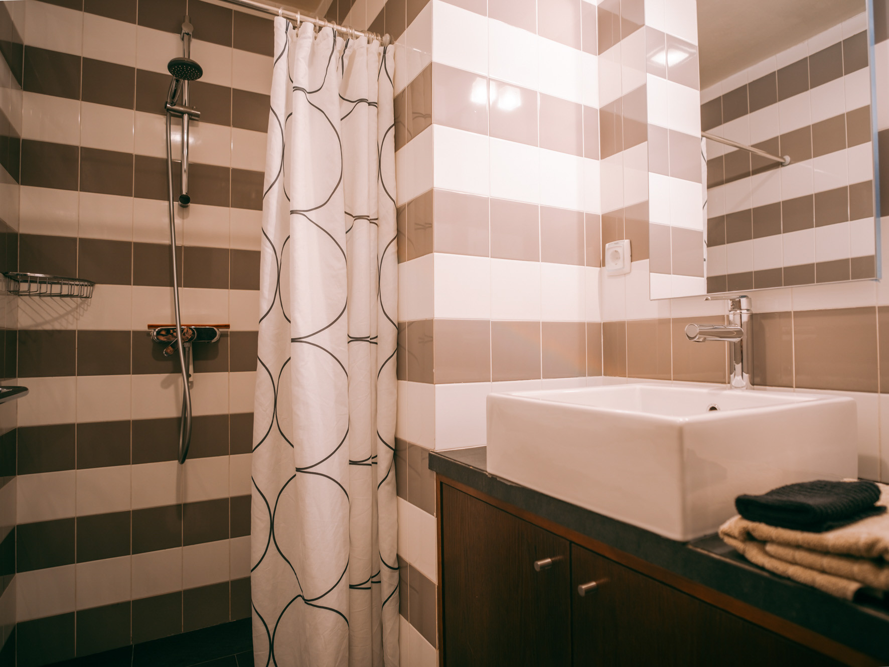 Bathroom with striped brown and white tiles, square sink on wooden cabinet, and shower with patterned curtain.