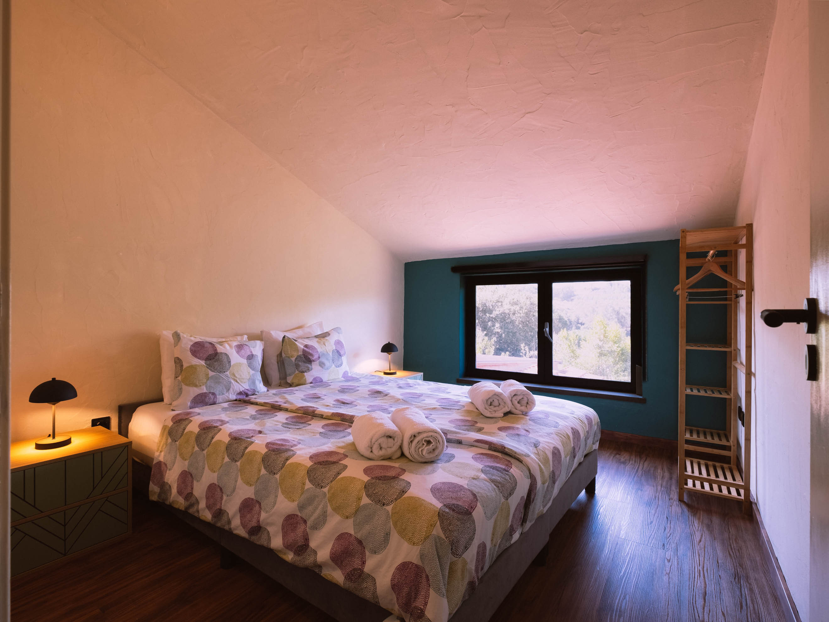 Bedroom with a double bed, patterned bedspread, rolled towels on bed, wooden floor, window, and clothes rack.