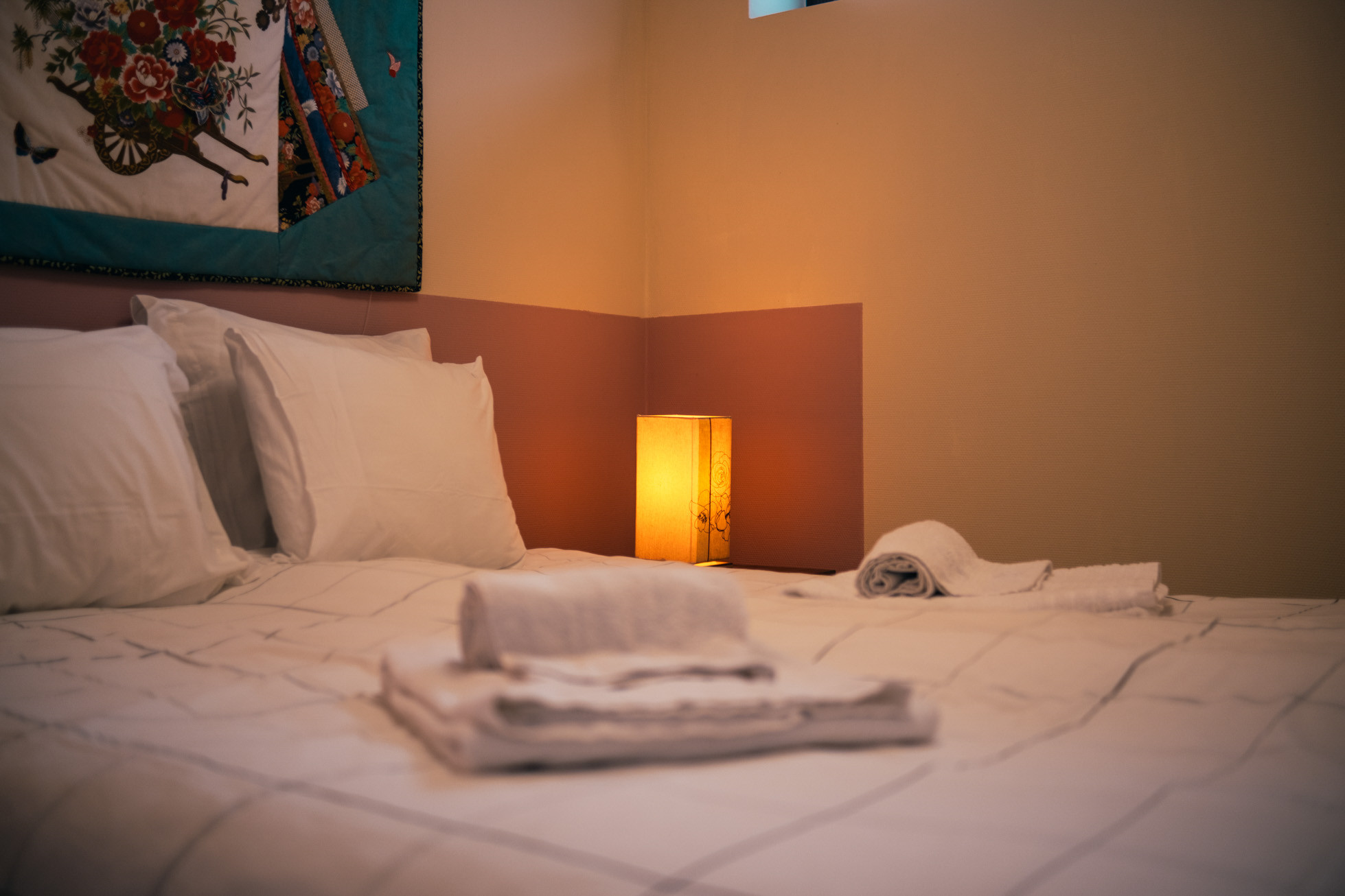 Bed with white linens and pillows, folded towels on top, lit bedside lamp and decorative wall hanging.