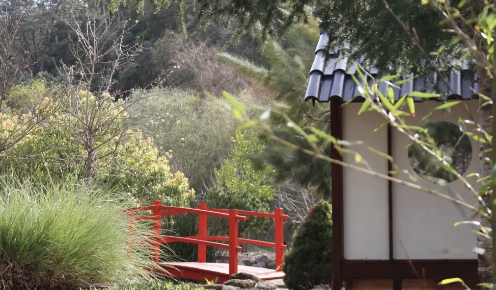 Red wooden garden bridge surrounded by greenery with part of a structure with tiled roof in foreground