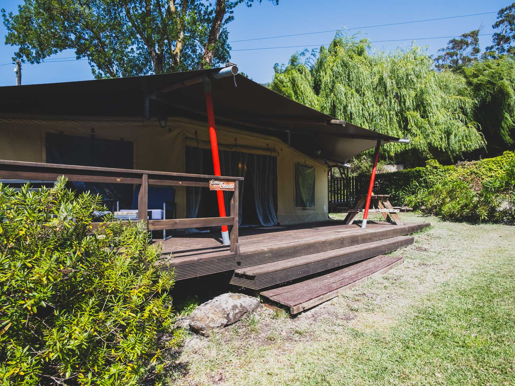 Wooden deck with steps and railing leads to a tent-like structure surrounded by green bushes and trees.