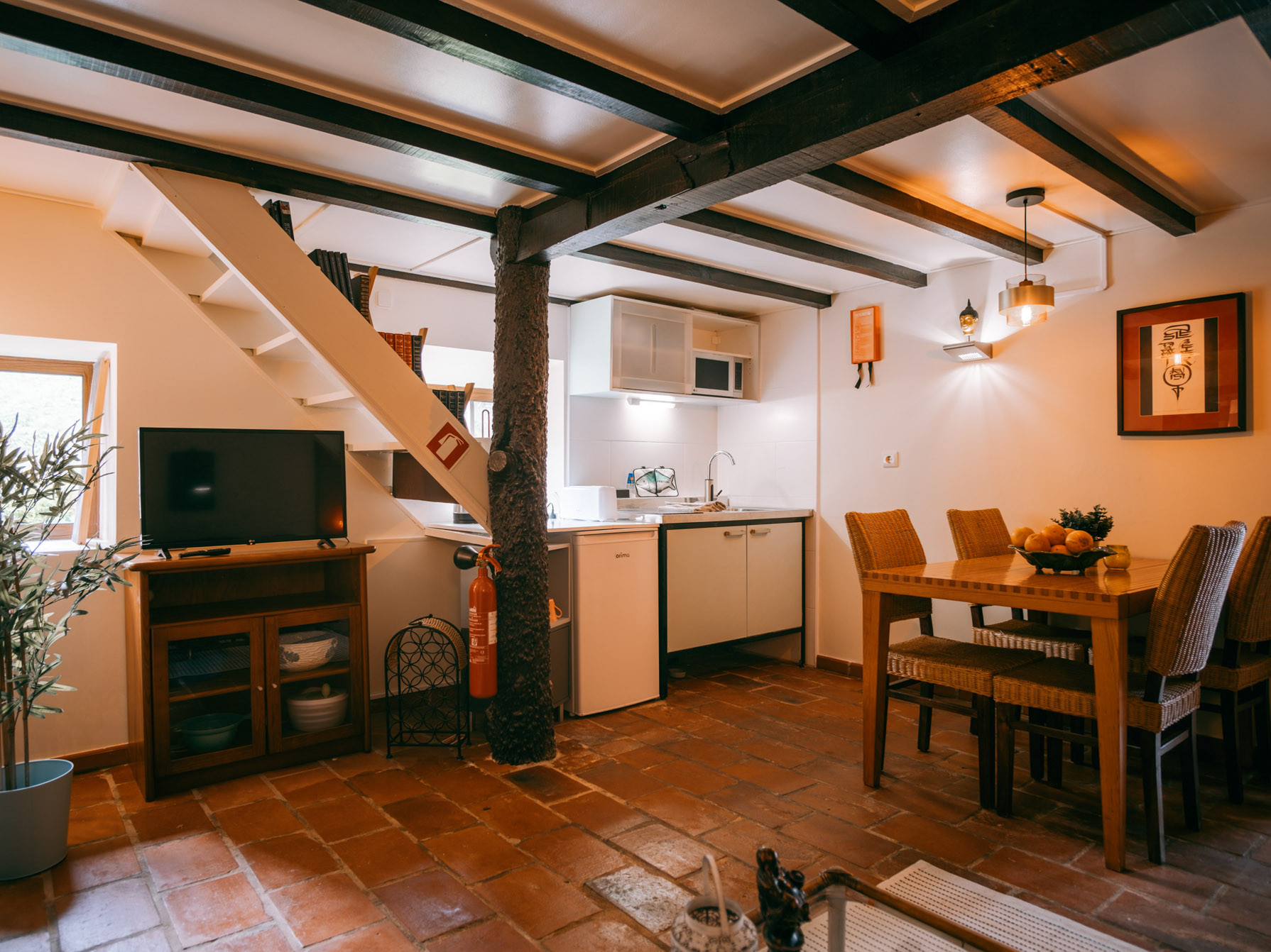 Cozy living space with kitchen, dining table, TV, staircase, and exposed ceiling beams.