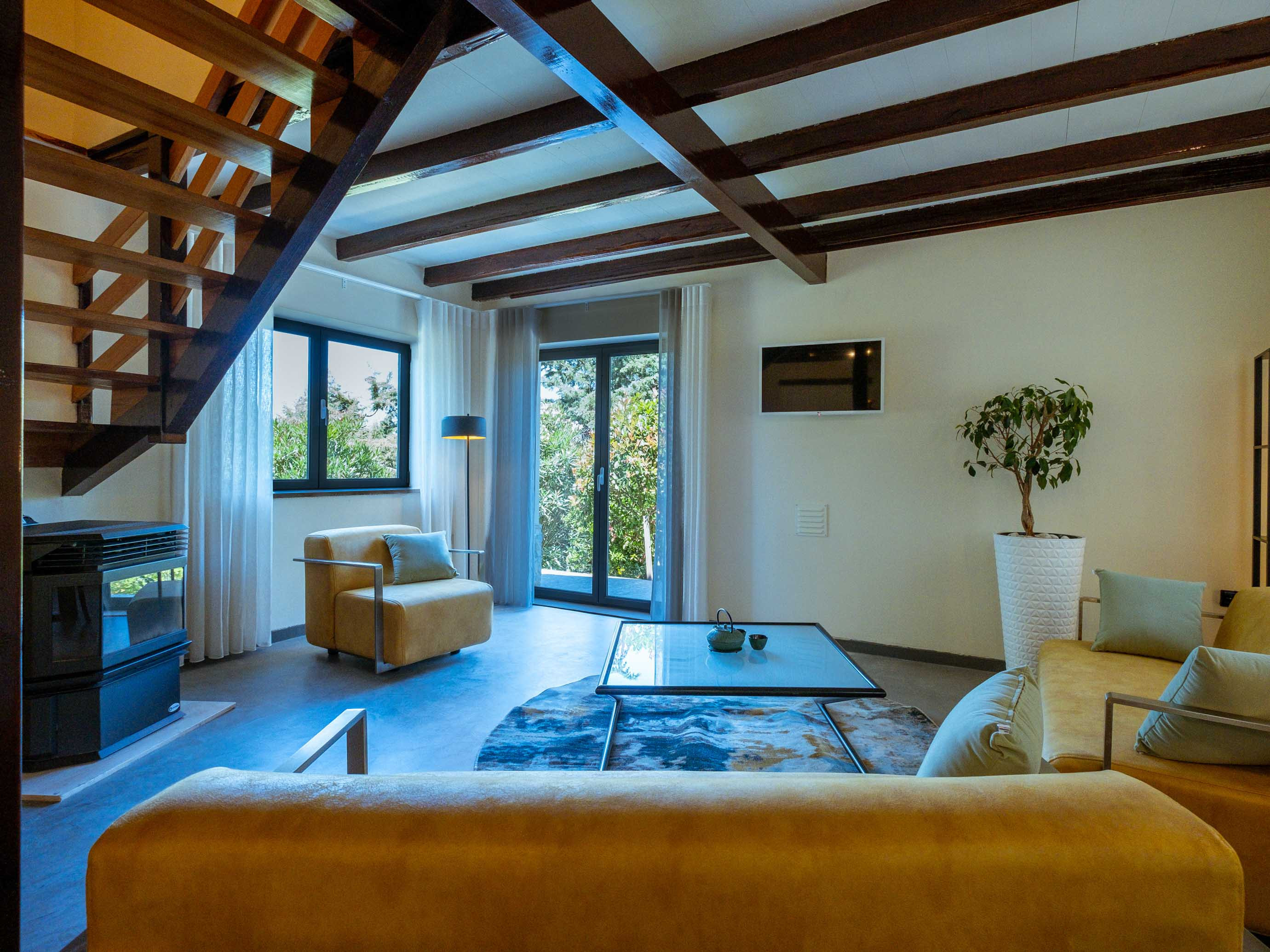 Living room with mustard yellow armchairs and sofa, glass coffee table, wooden ceiling beams, and large windows.