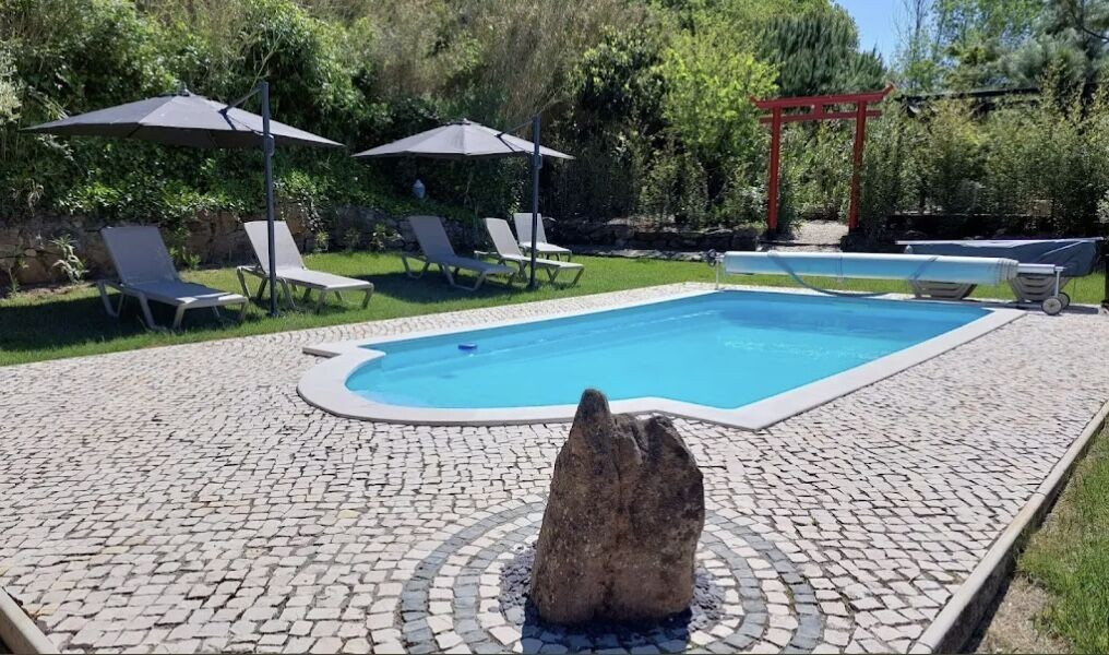 Outdoor pool with stone pavement, four lounge chairs under umbrellas, and a red Japanese-style gate in the background.