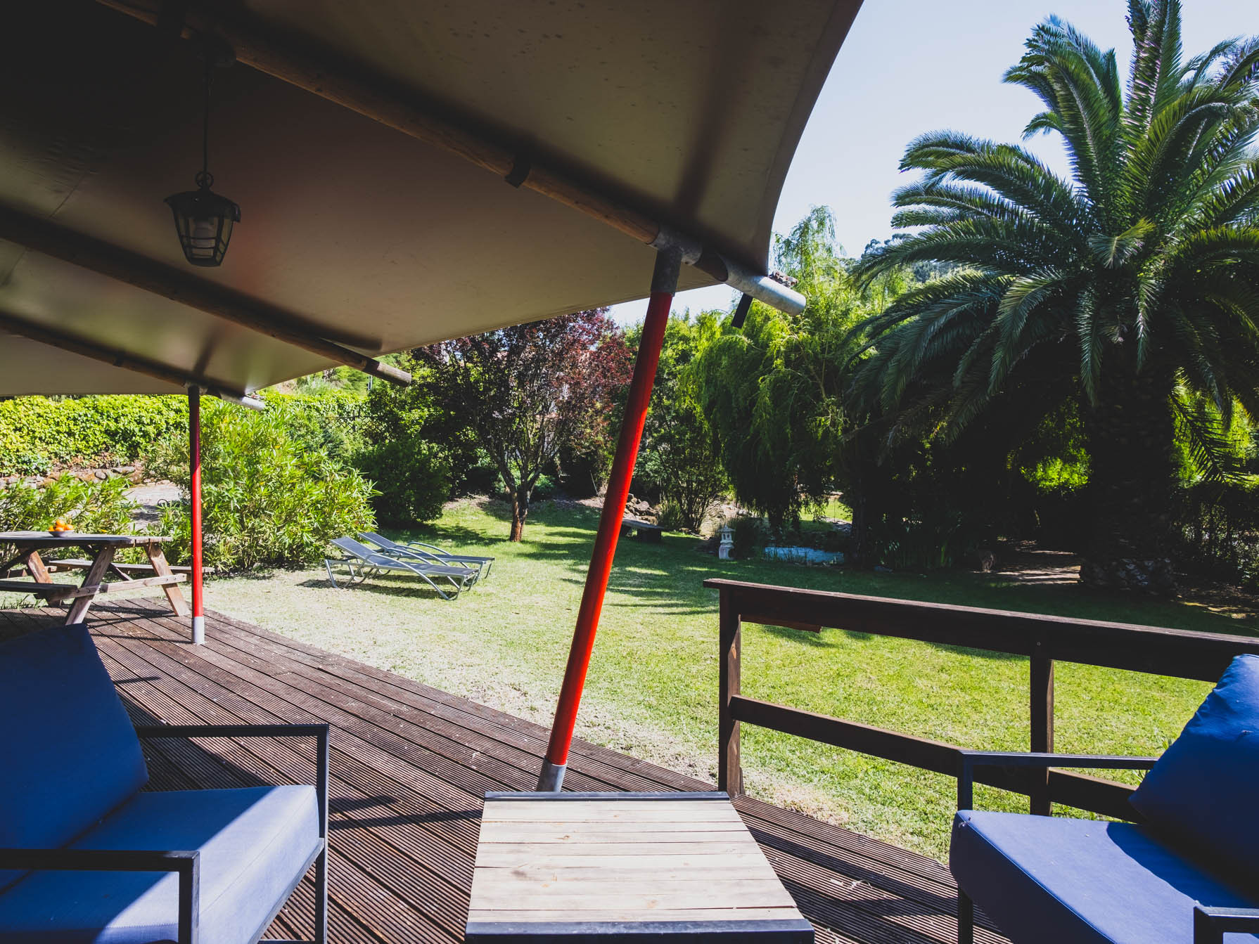 Covered wooden deck with blue cushioned chairs overlooking a garden with palm tree and lawn chairs.