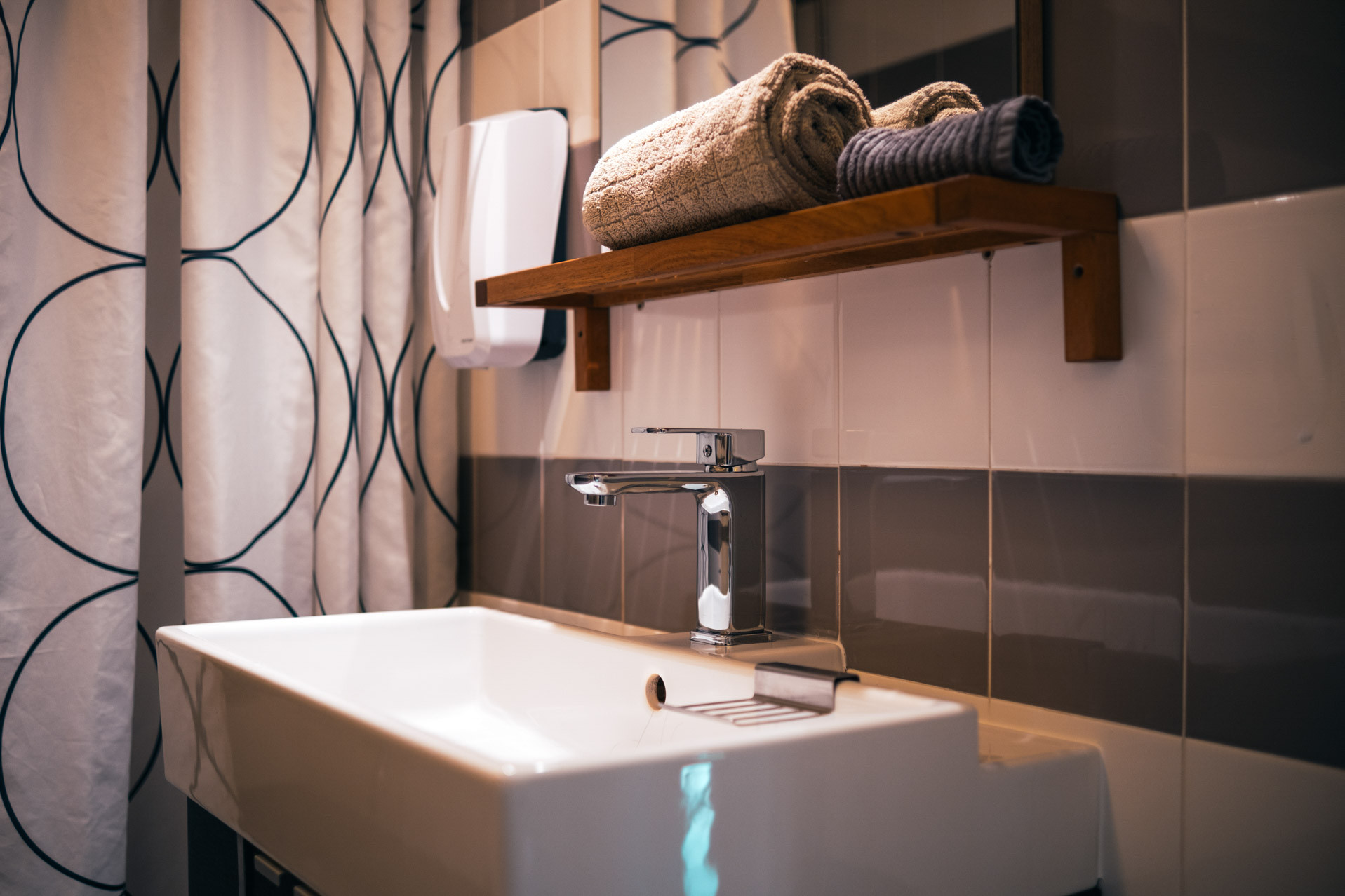 White rectangular sink with chrome faucet, wooden shelf above holding rolled towels and patterned shower curtain.