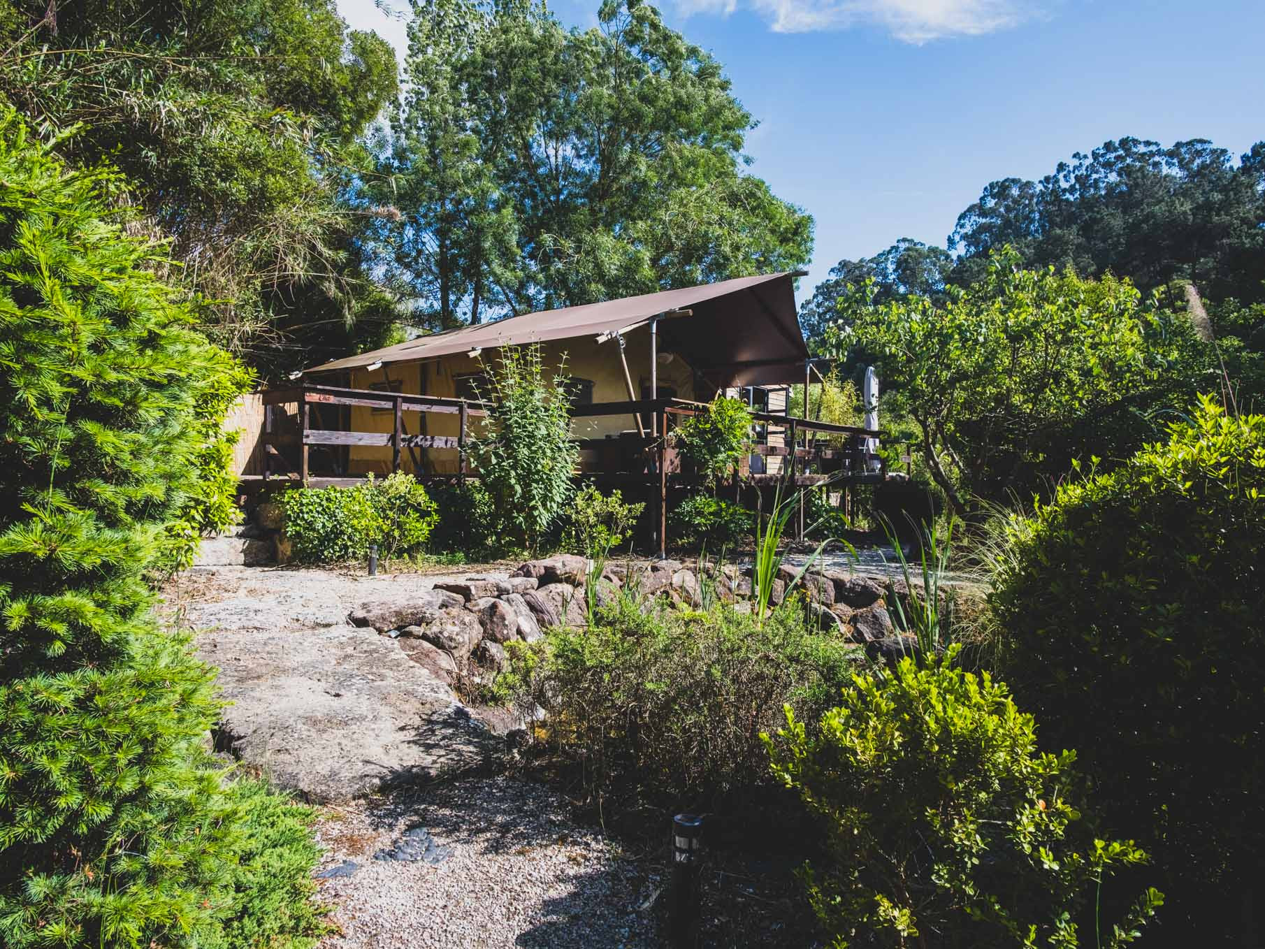 Raised tent structure with wooden railings surrounded by green trees and shrubs under a blue sky.