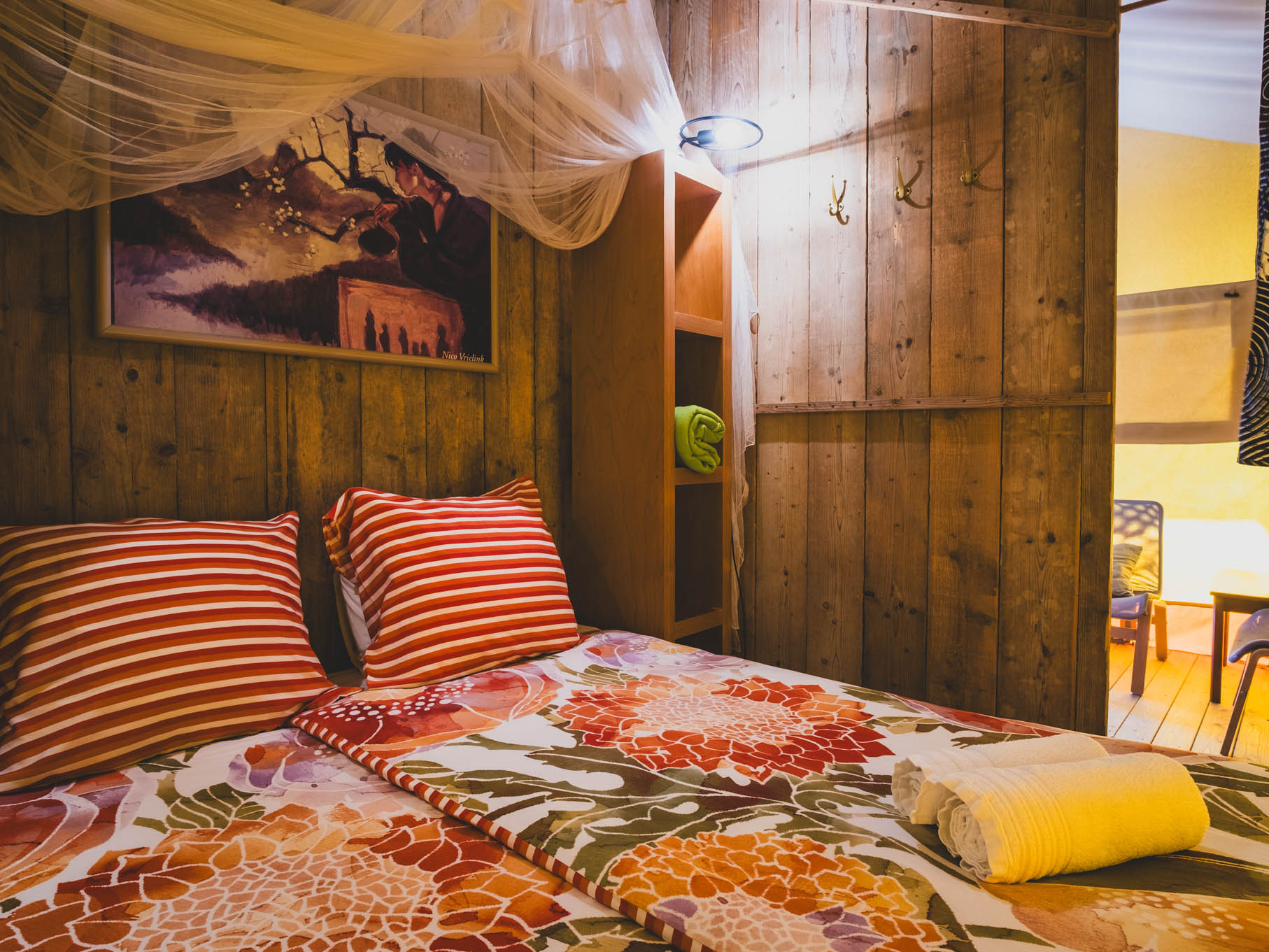 Bedroom with wooden walls, floral bedspread, striped pillows, rolled towels, and a painting above the bed.