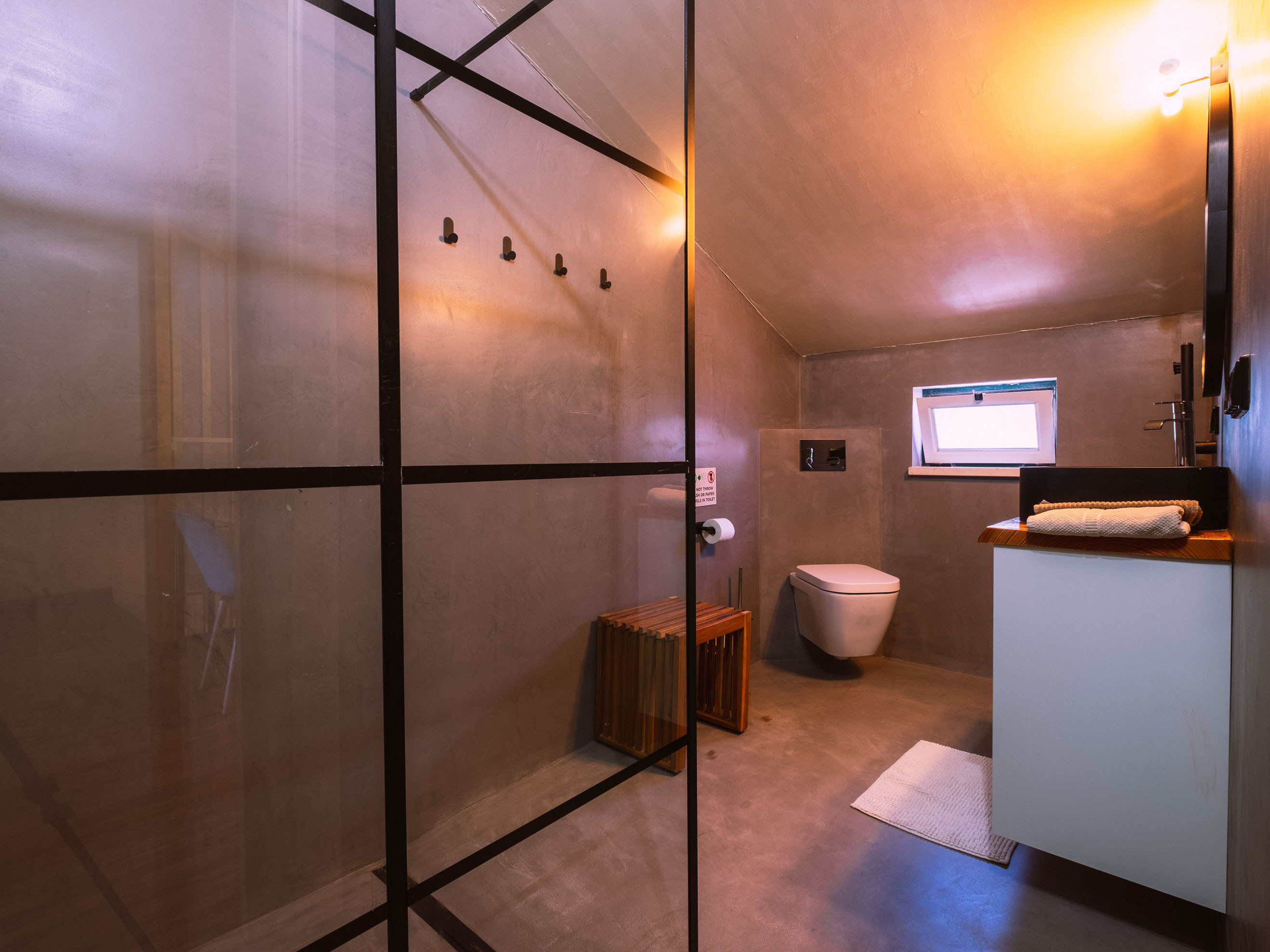 Bathroom with concrete walls, black-framed glass shower screen, wooden stool, and wall-mounted toilet under small window.