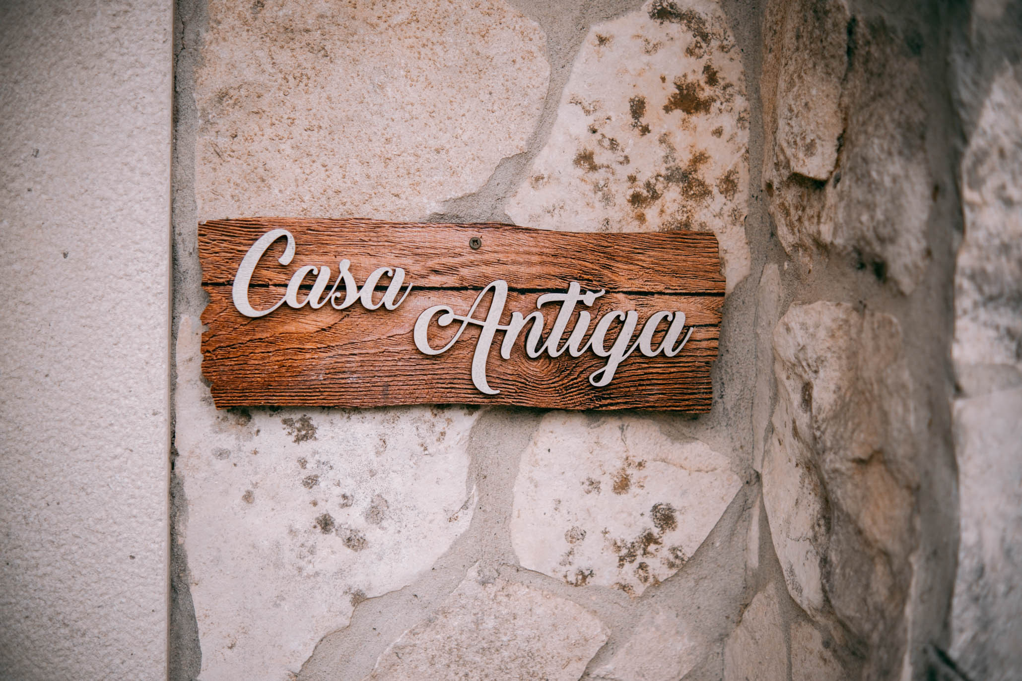 Wooden sign with white script letters reading Casa Antiga mounted on a stone wall.