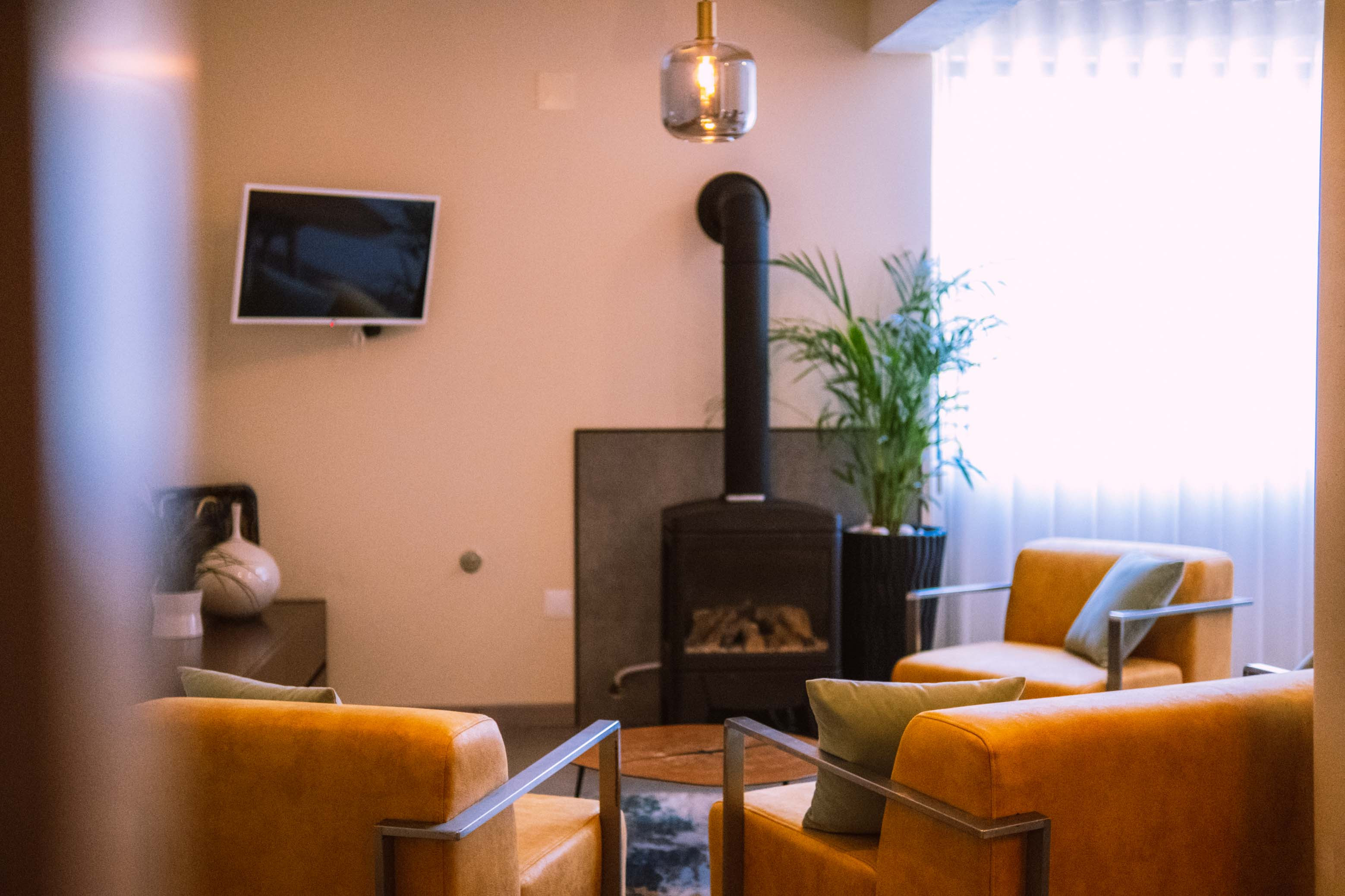 Cozy living room with mustard armchairs, wood stove, potted plant, wall TV, and hanging light fixture.
